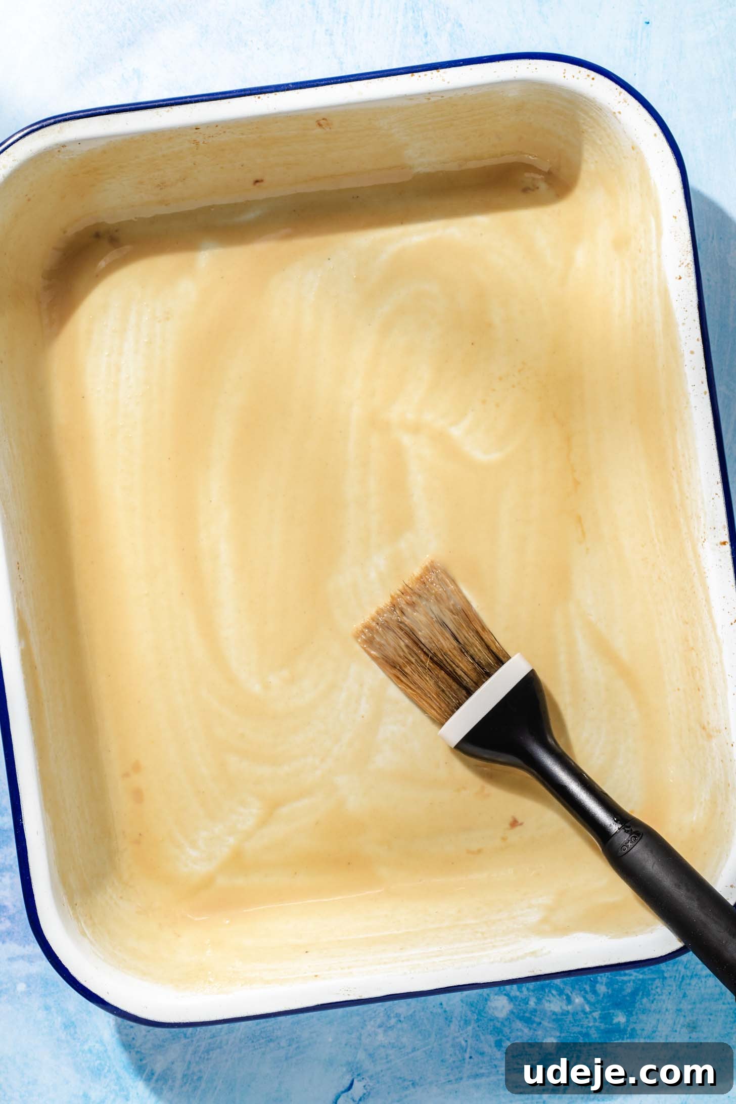 Aromatic Golden Semolina Cake 6 A white rectangular baking pan with a blue rim, thoroughly brushed with tahini, and a brush with a black handle resting beside it on a blue background.