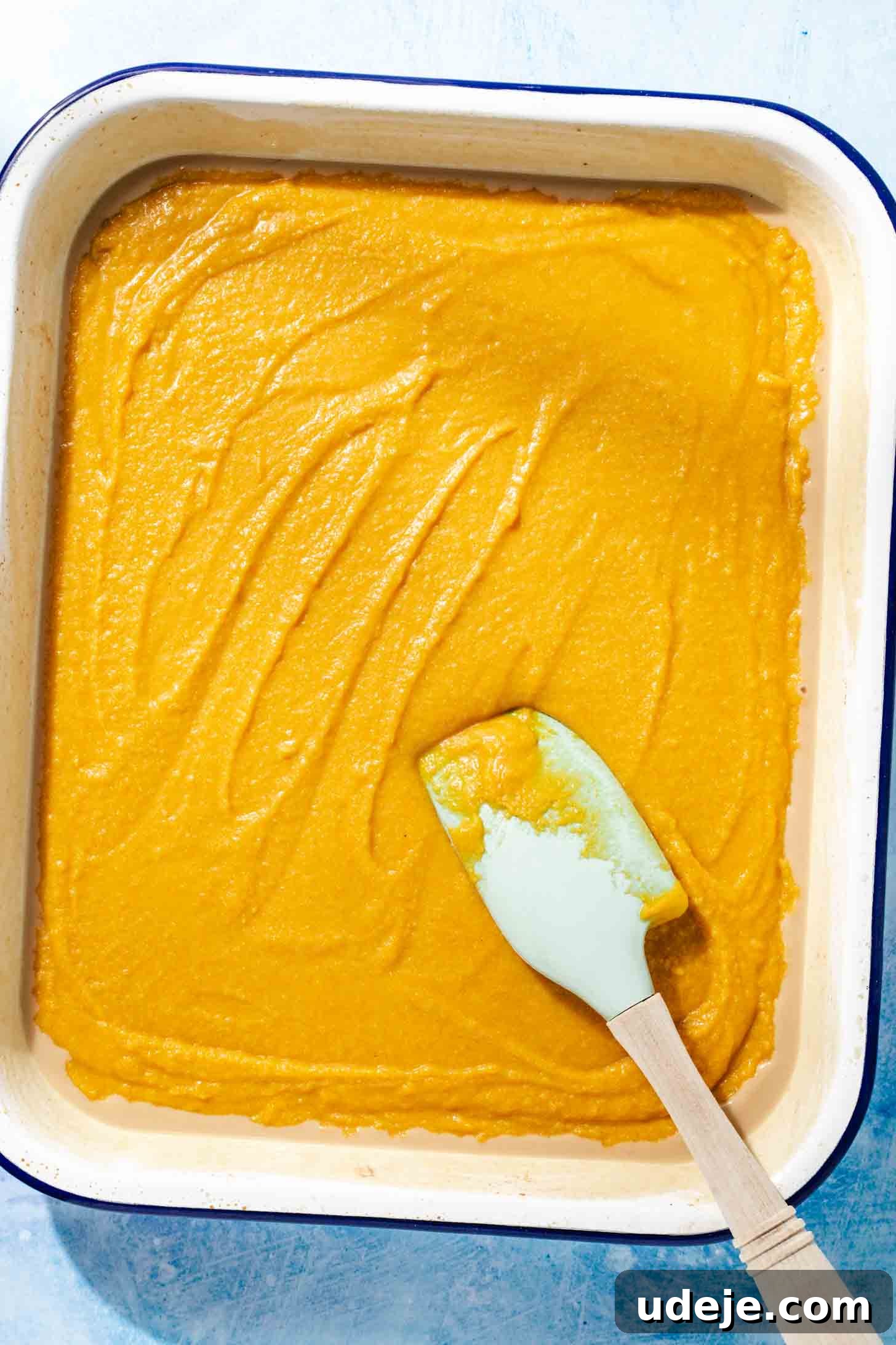 Aromatic Golden Semolina Cake 7 Yellow Sfouf batter just poured into a white rectangular baking pan with a blue rim, with a light blue spatula on top, all on a blue background.