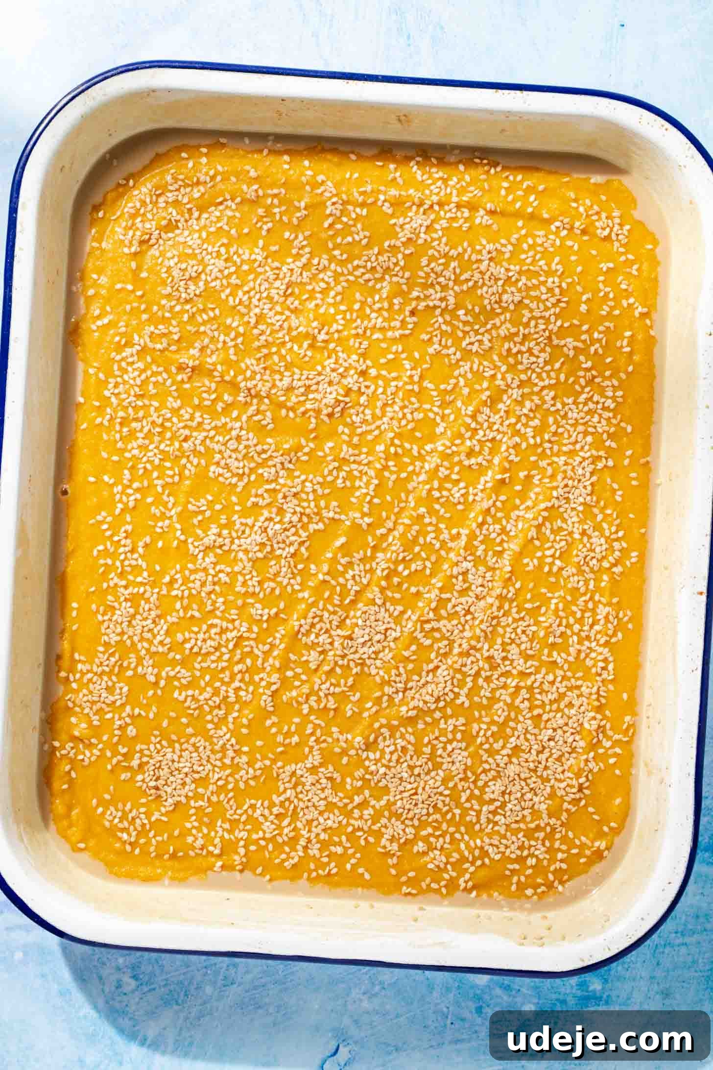 Aromatic Golden Semolina Cake 8 Yellow Sfouf batter in a white rectangular baking pan with a blue rim, generously sprinkled with toasted sesame seeds on top, all on a blue background.