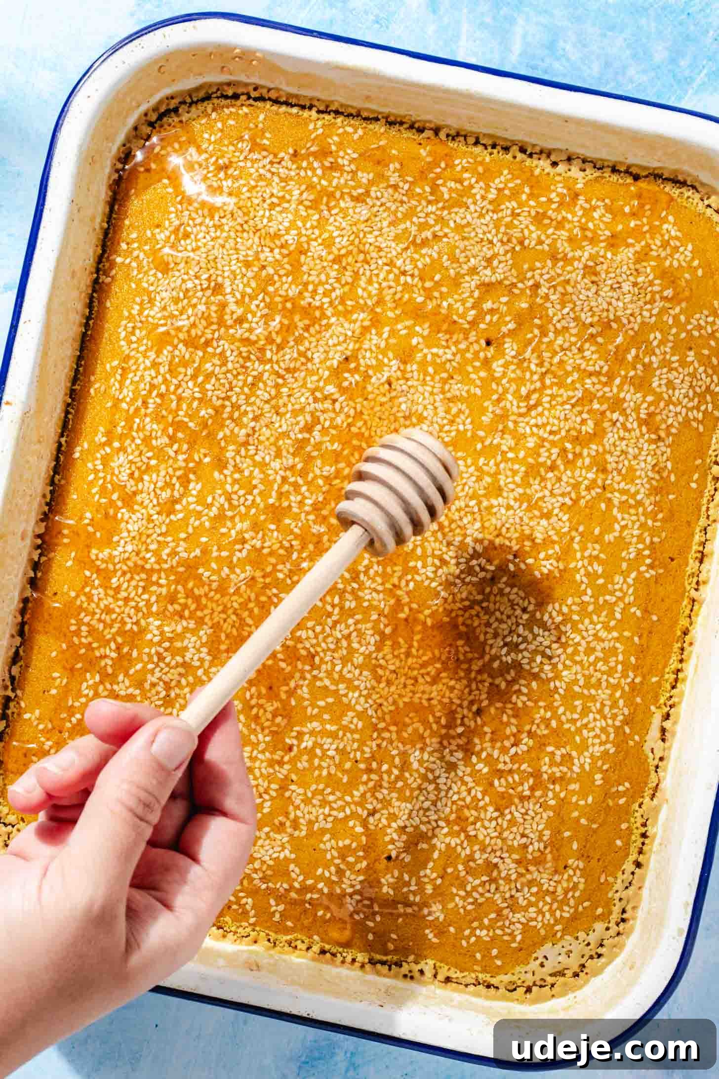 Aromatic Golden Semolina Cake 10 Freshly baked golden yellow Sfouf cake in a white rectangular baking pan with a blue rim, being drizzled with honey from a wooden honey dipper stick, all on a blue background.
