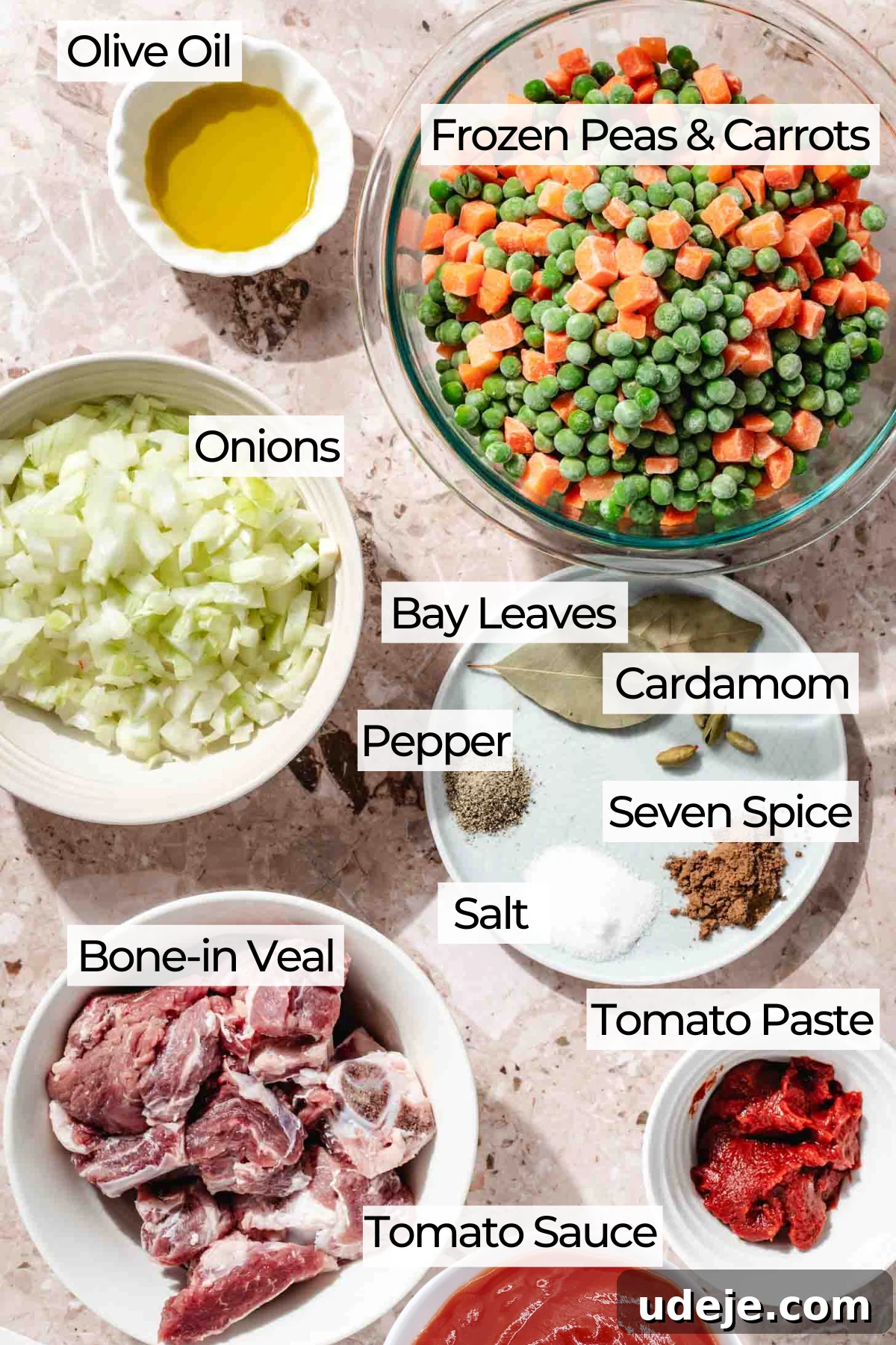 Ingredient shot with glass bowl of frozen peas and carrots, small white bowl with olive oil, white bowl with diced onions, plate with bay leaves, cardamom pods, seven spice, salt and pepper, white bowl with bone in veal, small white bowl with tomato paste, white bowl with tomato sauce on a beige marble background.