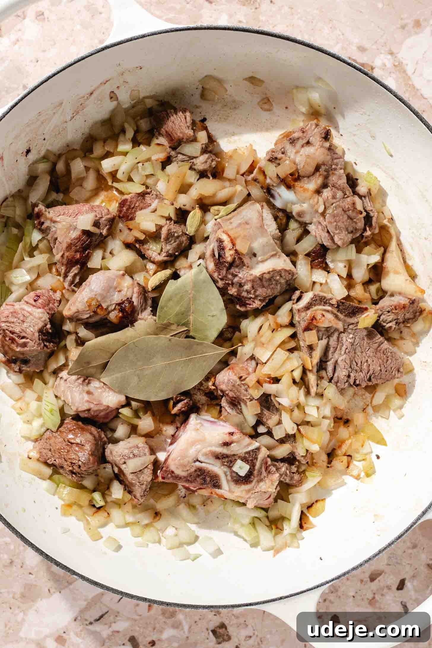 Bone-in veal with chopped onions sauteeing in a white pot with bay leaves on beige marble background.