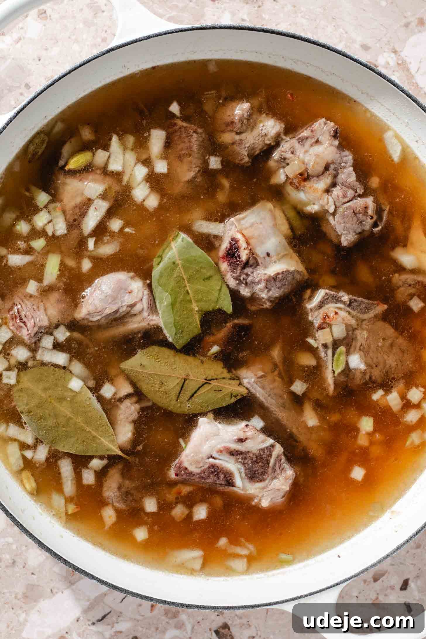 Bone-in veal with chopped onions, bay leaves and water in a white pot on a beige marble background.
