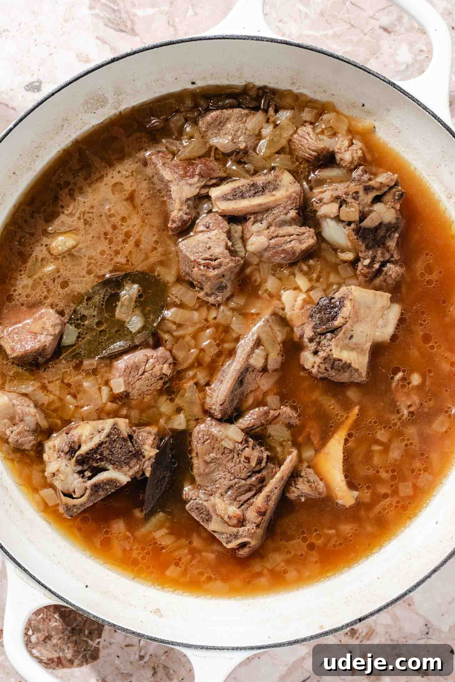 Bone-in veal with chopped onions, bay leaves and water in a white pot on a beige marble background.