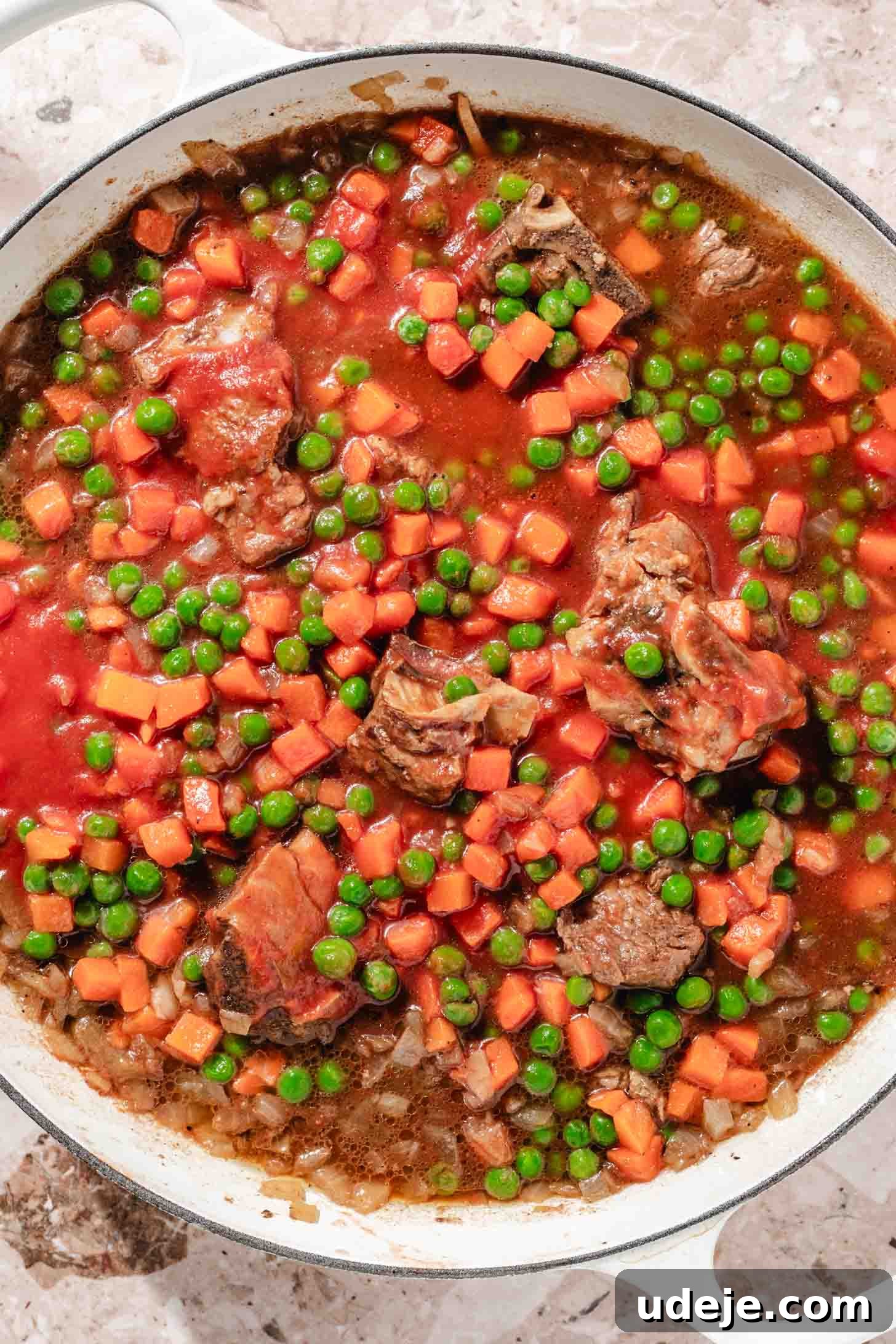 Veal with a tomato base and frozen peas and carrots in a white pot on a beige marble background.