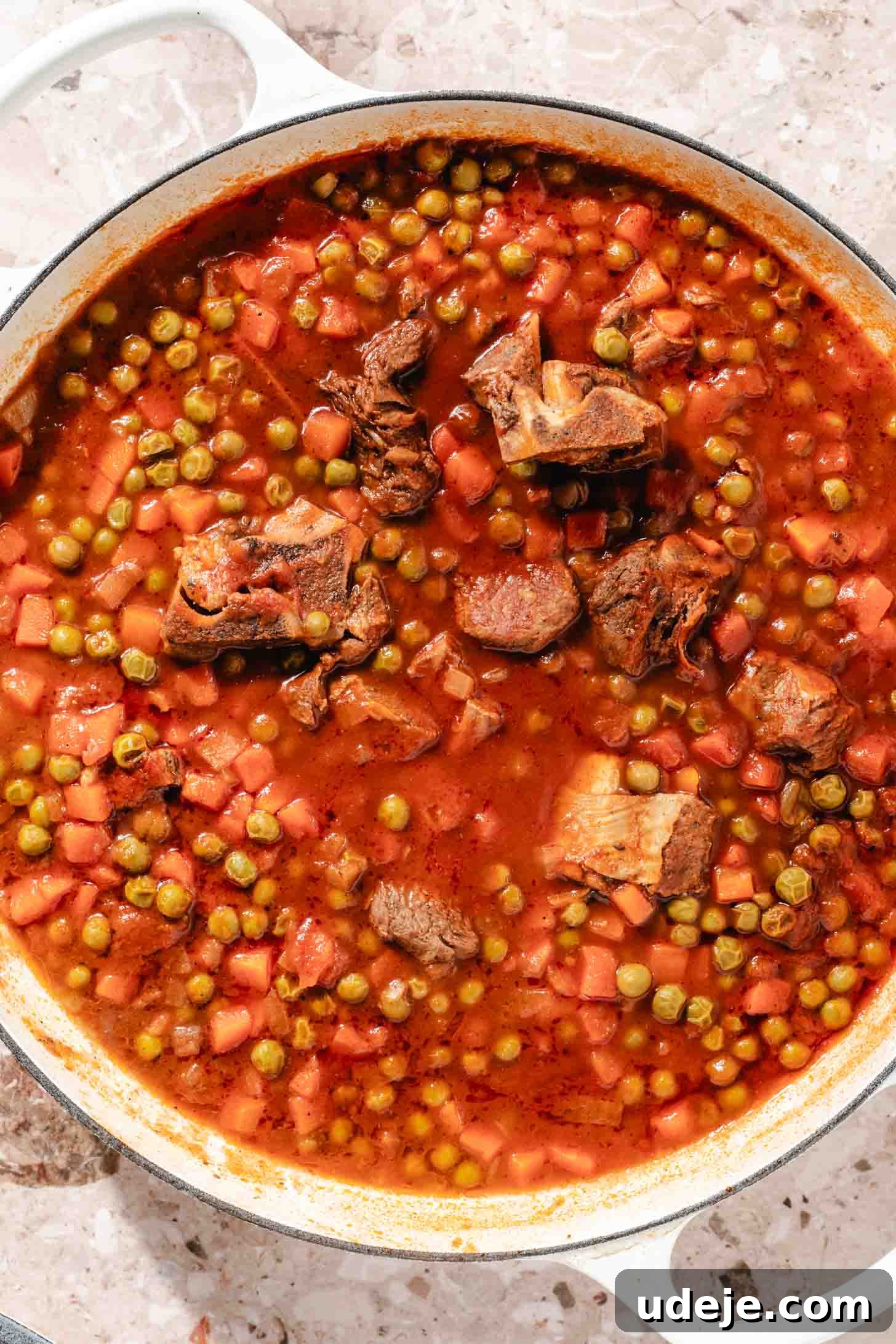 Veal with a tomato base and frozen peas and carrots in a white pot on a beige marble background.