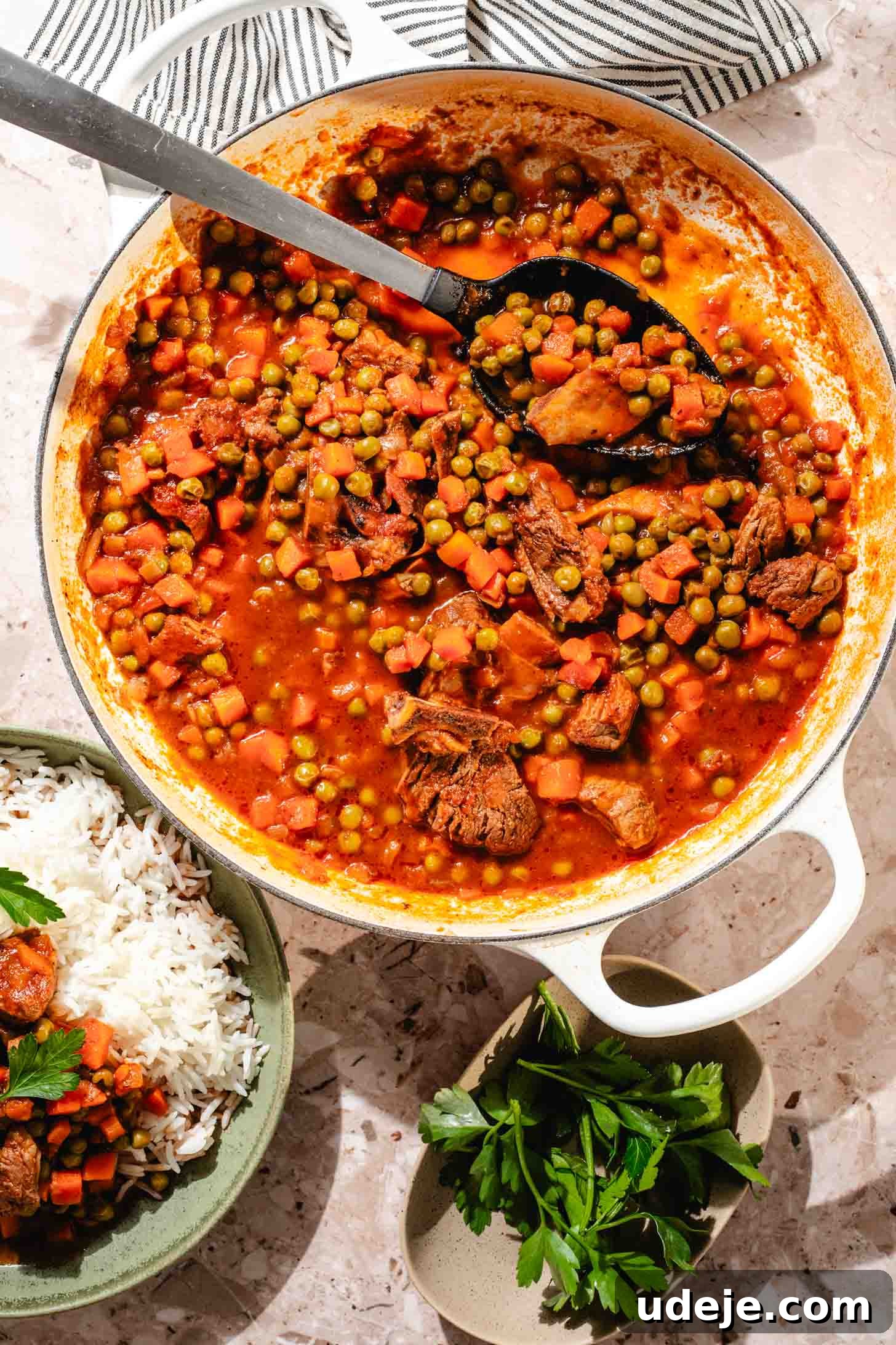 Veal stew with peas and carrots in a white pot with a black and metal serving spoon with a green bowl with white rice and stew on top, a small bowl of parsley, a black and white striped tea towel on a beige marble background.