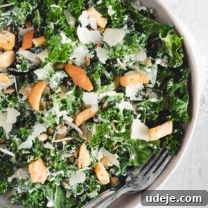 A bowl of kale tahini salad Caesar-style with croutons, sunflower seeds, shaved Parmesan cheese, and creamy dressing, served with a fork on a white surface.
