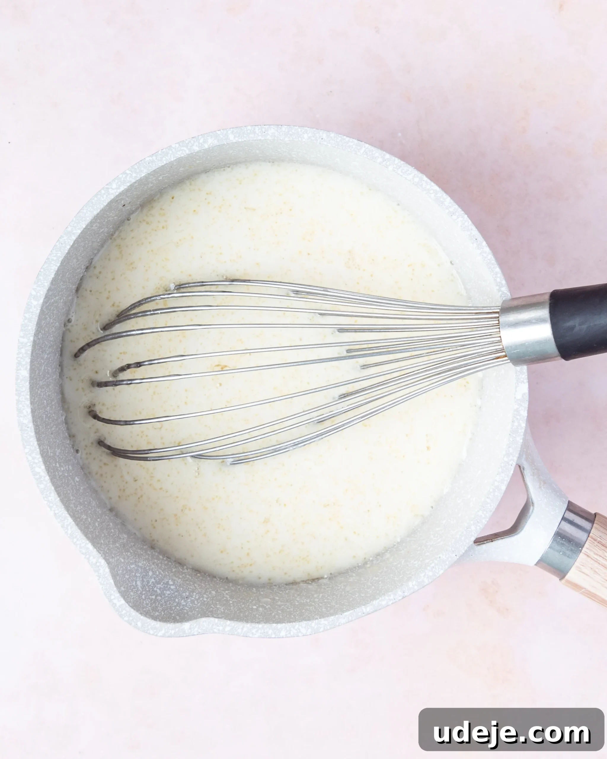 Homemade Turkish Delight: Customize with Pistachios 11 The cornstarch liquid thoroughly mixed with the sugar syrup, now a combined mixture in a small light grey saucepan, being continuously whisked on a soft pink background.