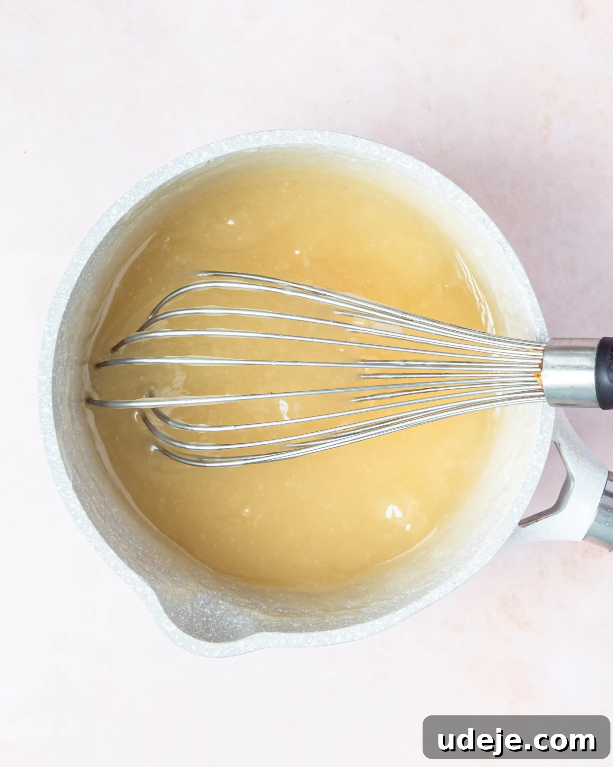 Homemade Turkish Delight: Customize with Pistachios 12 A light brown, gelled mixture inside a small light grey saucepan, showing its thickening consistency with a whisk resting in it, on a soft pink background.