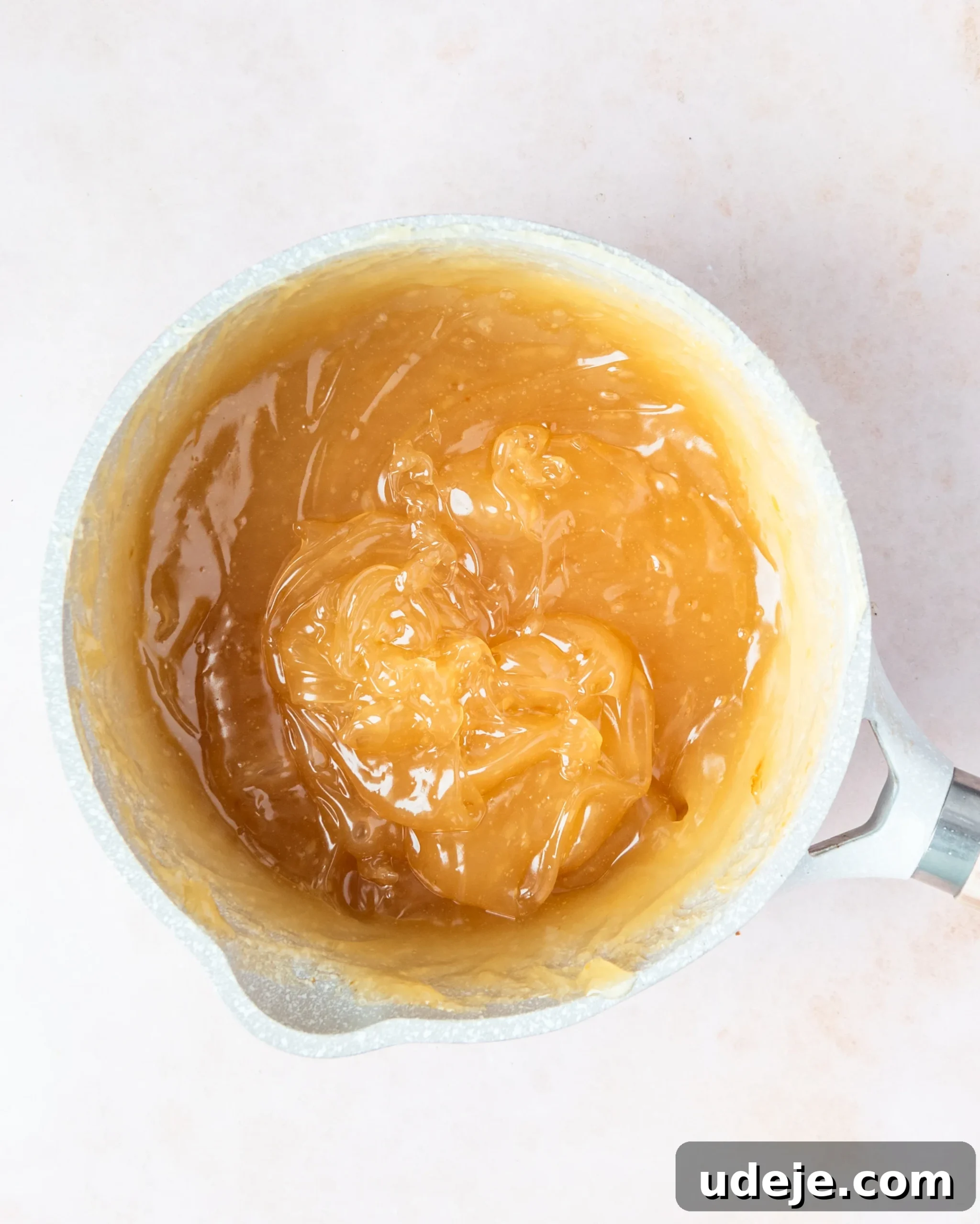 Homemade Turkish Delight: Customize with Pistachios 13 A medium brown, glossy, gelled mixture now completely thickened in a small light grey saucepan, ready for the next steps, on a soft pink background.