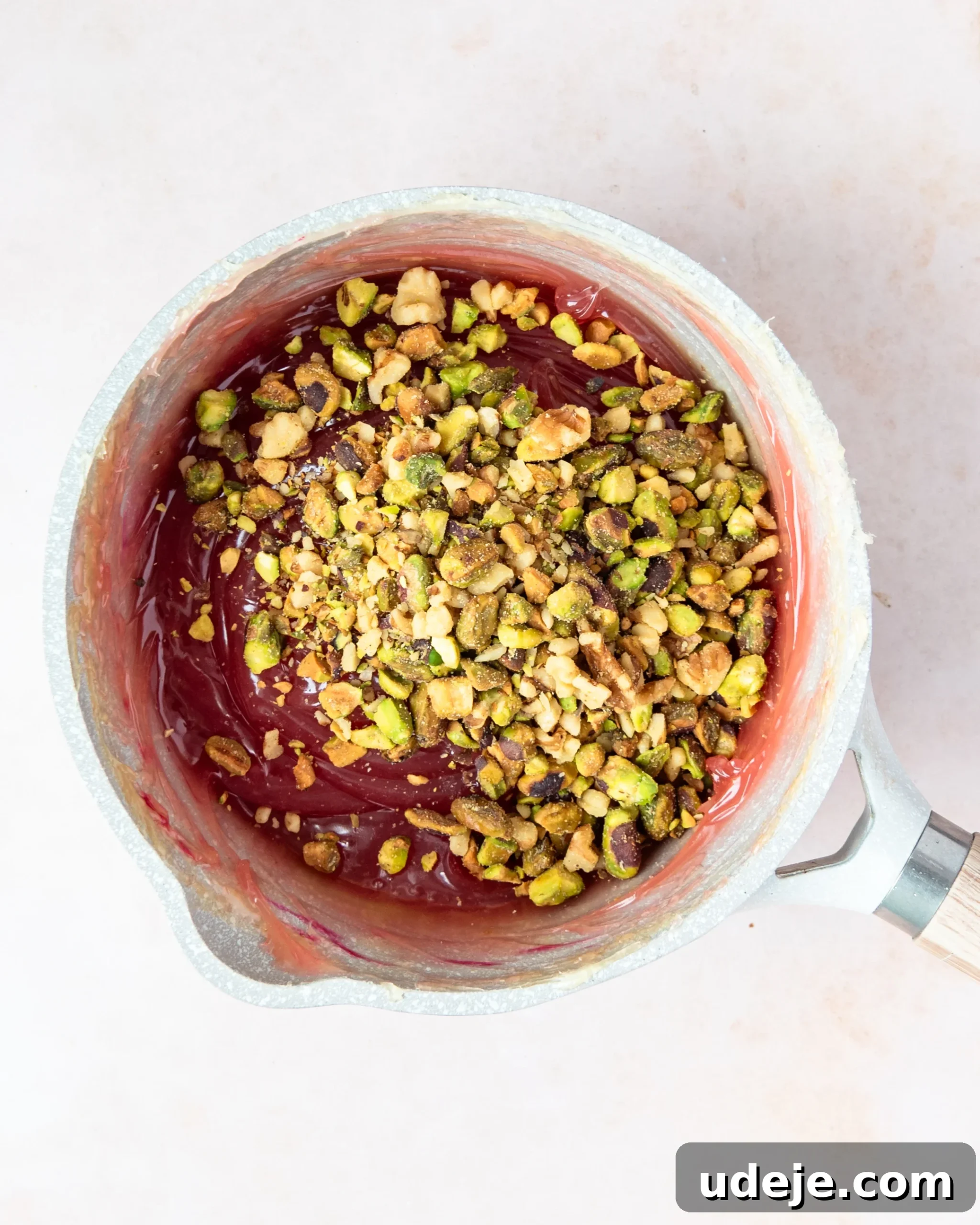 Homemade Turkish Delight: Customize with Pistachios 15 A gelled red mixture in a small light grey saucepan, now with chopped pistachios sprinkled on top, ready to be mixed in, on a soft pink background.
