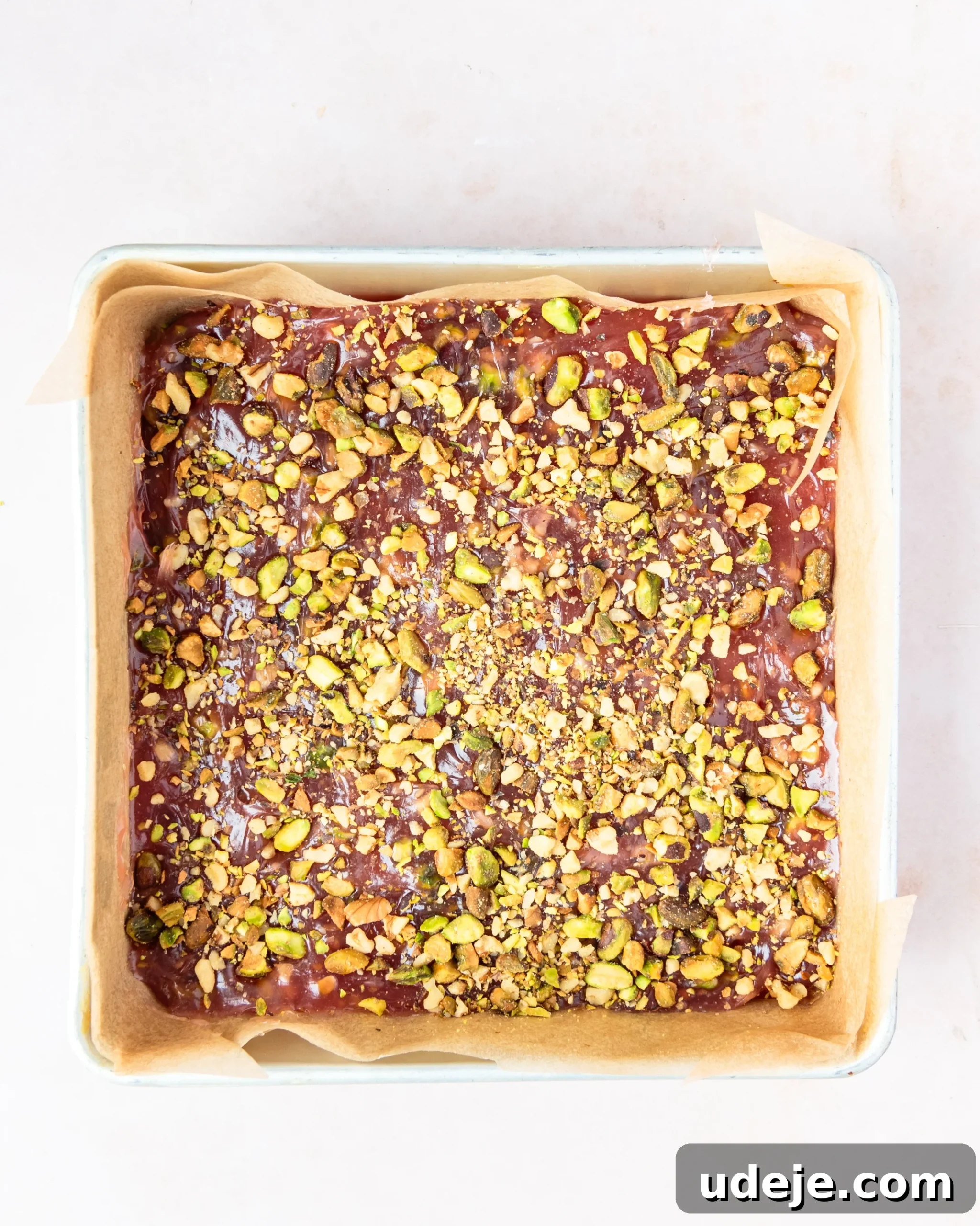 Homemade Turkish Delight: Customize with Pistachios 17 The red gelled mixture, generously mixed with chopped pistachios, now carefully poured and smoothed into a parchment paper-lined square metal pan, on a soft pink background.