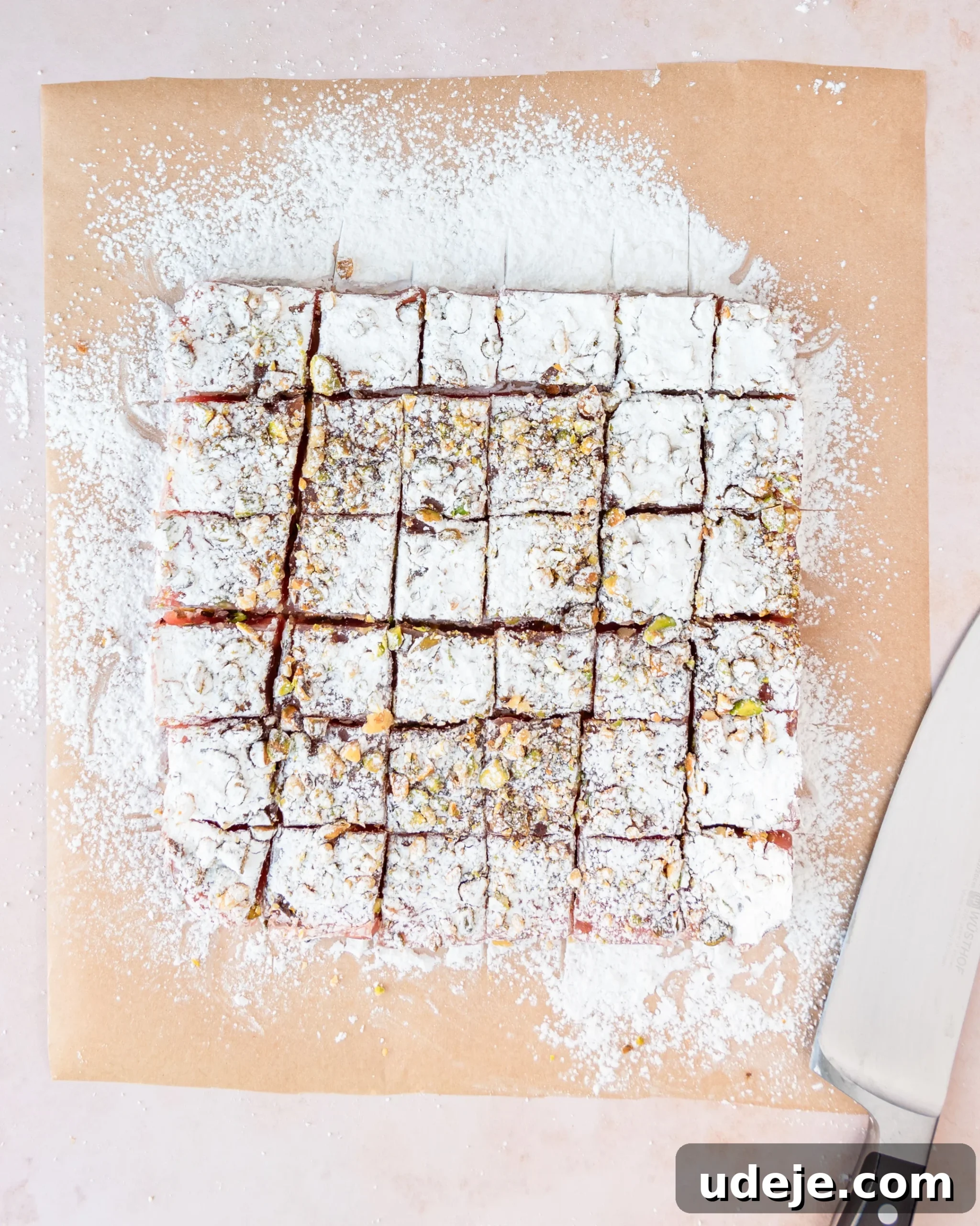 Homemade Turkish Delight: Customize with Pistachios 21 The set Turkish Delight block, dusted with powdered sugar, now expertly cut into precise squares using an oiled knife, ready for final coating, on a light pink background.