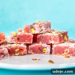 Squares of turkish delight stacked on a white plate against an aqua blue background, garnished with pistachios.