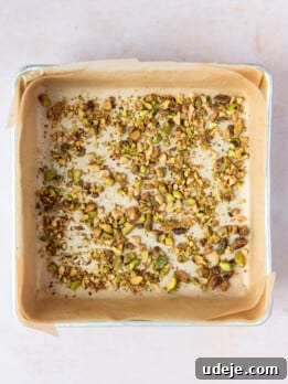 A metal square pan lined with light brown parchment paper with chopped pistachios on a soft pink background.