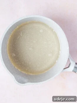Sugar syrup in a small light grey saucepan on a soft pink background.