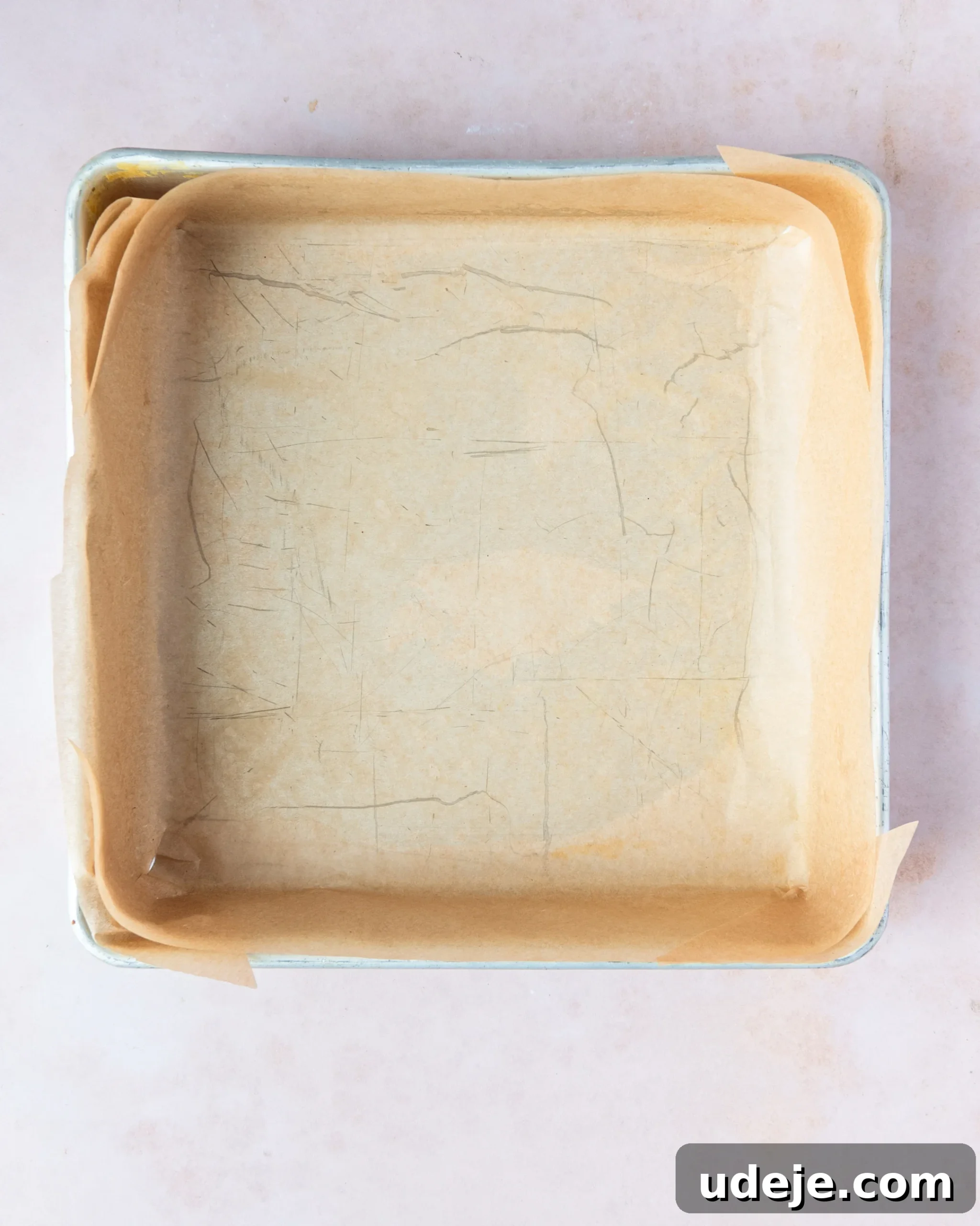 Homemade Turkish Delight: Customize with Pistachios 4 A metal square pan meticulously lined with light brown parchment paper, ready for the Turkish Delight mixture, presented on a soft pink background.