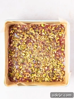 Gelled red mixture mixed with chopped pistachios in a parchment paper lined square metal pan on a soft pink background.