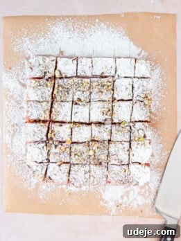 Powdered sugar on a piece of light brown parchment paper with a knife at the side with the set turkish delight on top with powdered sugar dusted on top and cut into squares on a light pink background.