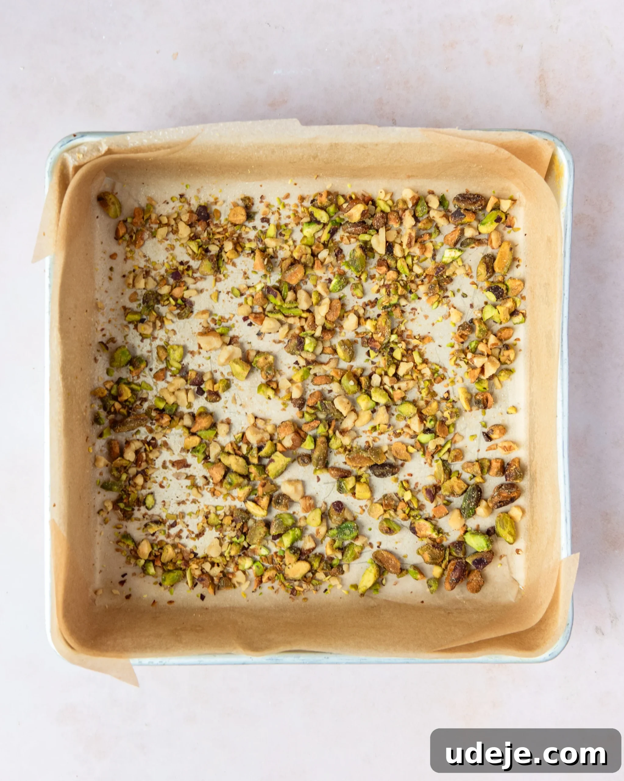 Homemade Turkish Delight: Customize with Pistachios 5 A metal square pan, perfectly lined with light brown parchment paper and generously sprinkled with chopped pistachios, resting on a soft pink background.