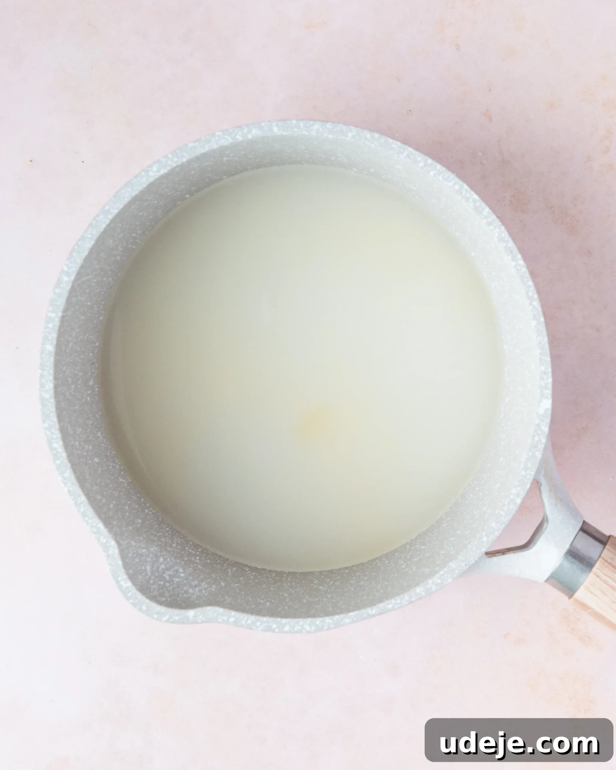 Homemade Turkish Delight: Customize with Pistachios 6 A light grey saucepan containing a mixture of water, sugar, and lemon juice, simmering gently on a soft pink background.