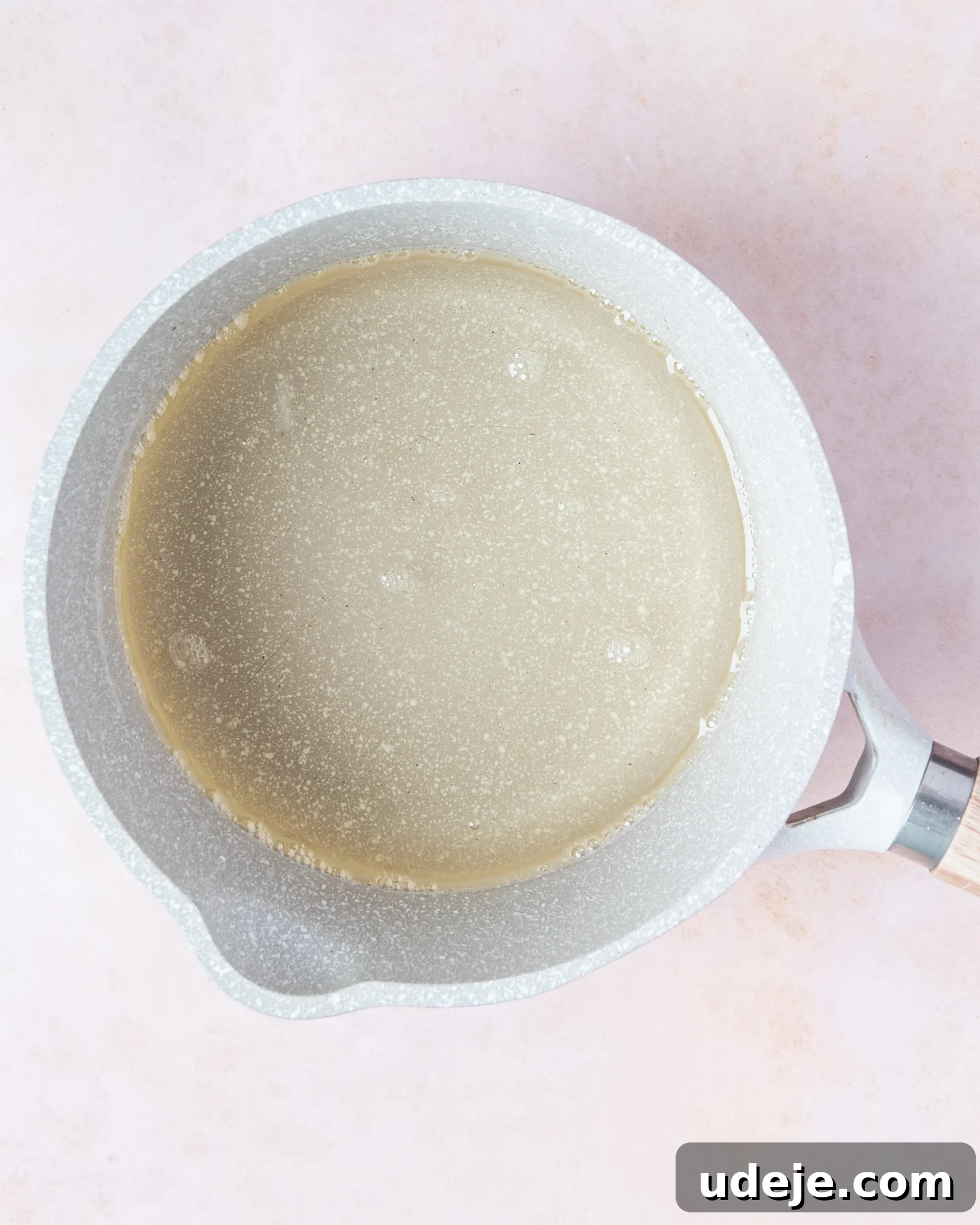 Homemade Turkish Delight: Customize with Pistachios 7 A glistening sugar syrup mixture in a small light grey saucepan, having thickened and become glossy after simmering, on a soft pink background.