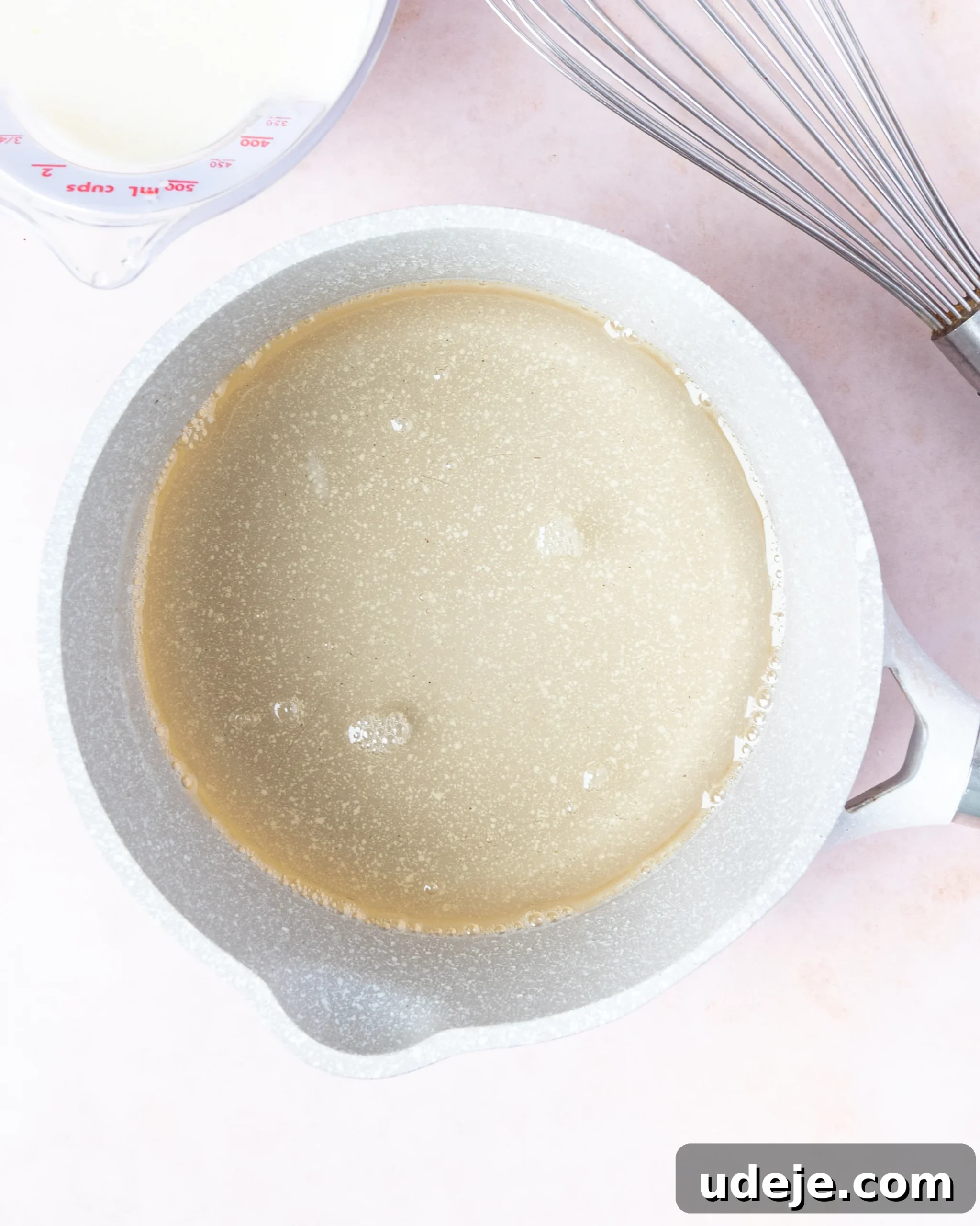 Homemade Turkish Delight: Customize with Pistachios 10 The process of combining sugar syrup in a light grey saucepan with the white liquid cornstarch mixture from a measuring cup, with a metal whisk ready for stirring, on a soft pink backdrop.