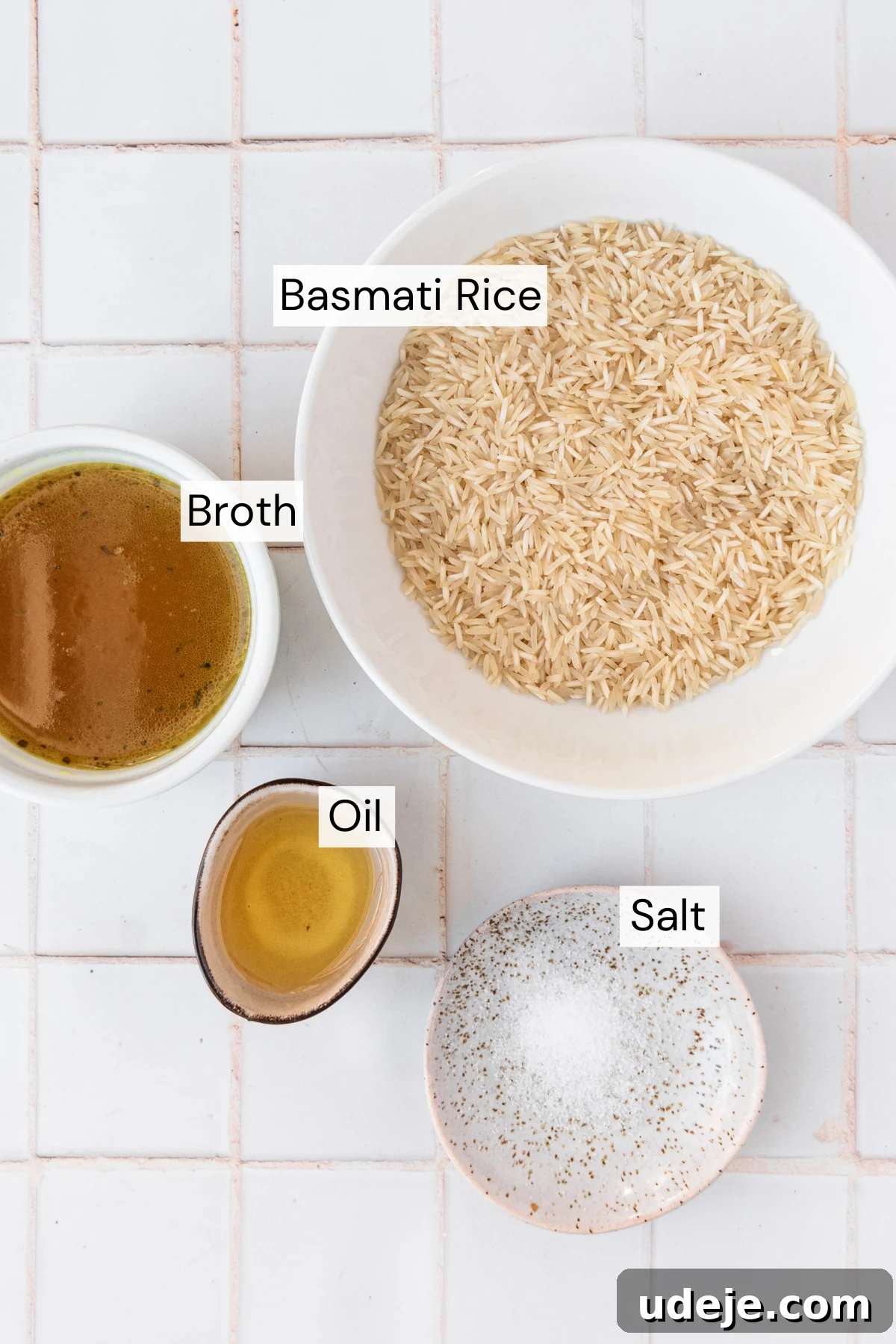 Instant Pot Basmati: Fluffy Perfection in 15 Minutes 3 A beautifully arranged ingredient shot featuring a white bowl of unwashed basmati rice, a small white dish of clear hot broth, a small brown bowl of oil, and a small speckled plate of salt, all set on a pristine white tiled background. This image highlights the fresh components needed for perfect Instant Pot Basmati Rice.