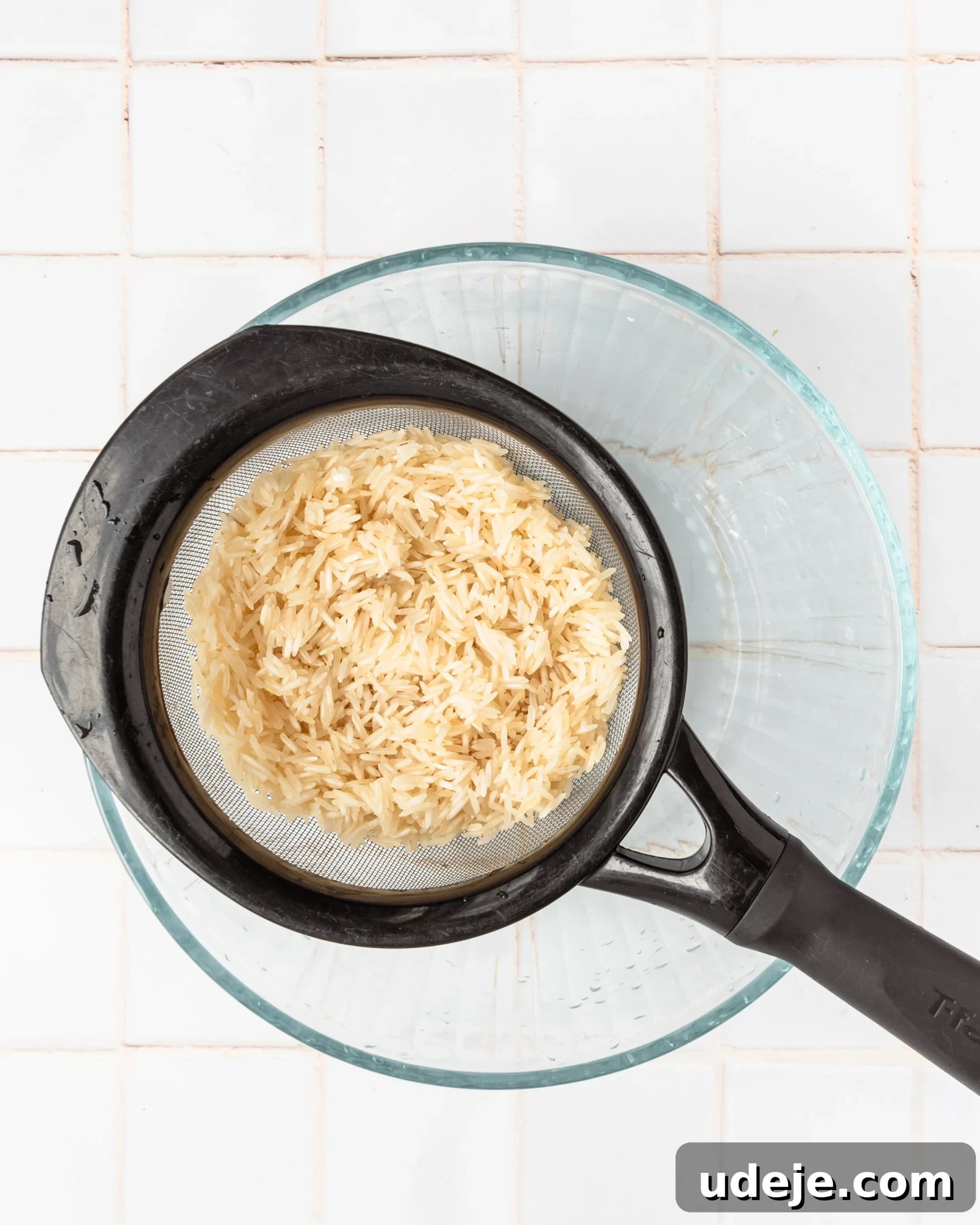 Instant Pot Basmati: Fluffy Perfection in 15 Minutes 6 Basmati rice thoroughly drained in a strainer, positioned over a glass bowl to catch any remaining water, set on a white tiled background. This step ensures excess moisture is removed before cooking.