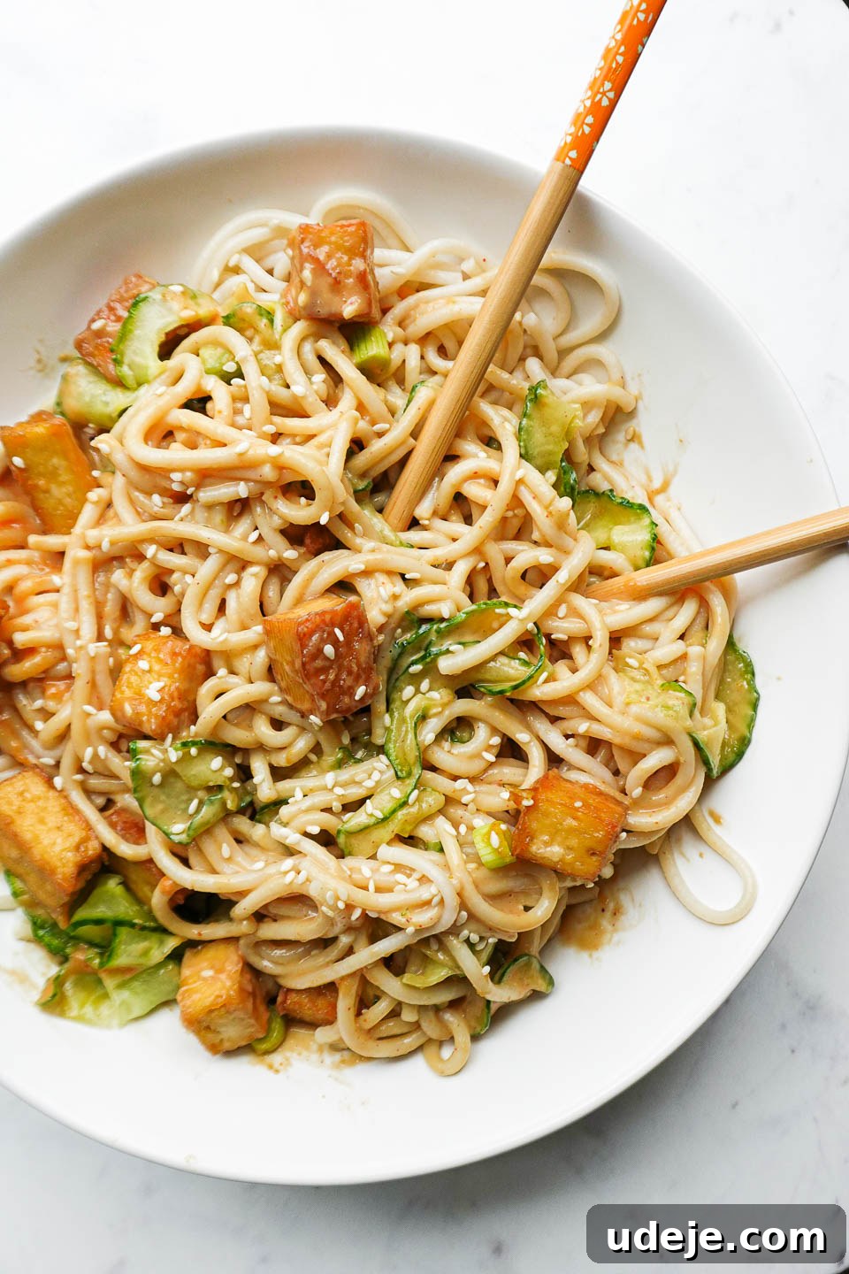 A vibrant bowl of peanut noodles featuring crispy tofu cubes, thin strands of spiralized cucumber, chopped green scallions, and a sprinkle of sesame seeds, presented with chopsticks.