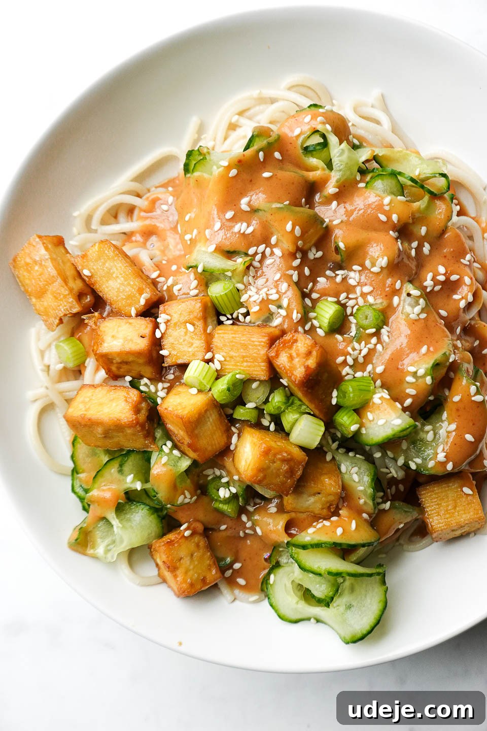A top-down view of vegan peanut noodles with crispy tofu and fresh vegetables served in a clean white bowl on a elegant marble surface.