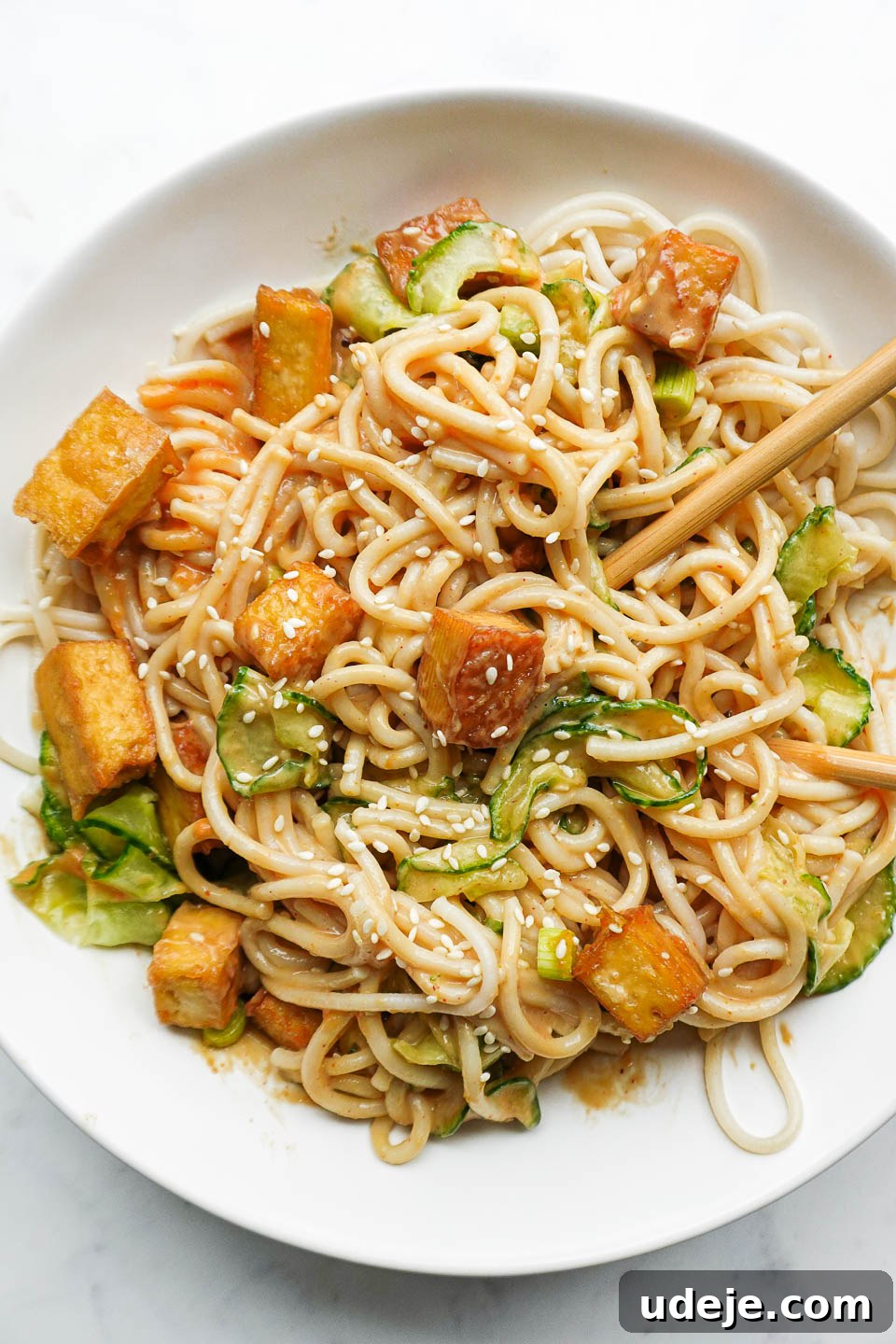 A close-up of a serving of peanut noodles, artfully garnished with crispy tofu, delicate spiralized cucumber, vibrant green scallions, and toasted sesame seeds, ready to be enjoyed with chopsticks.