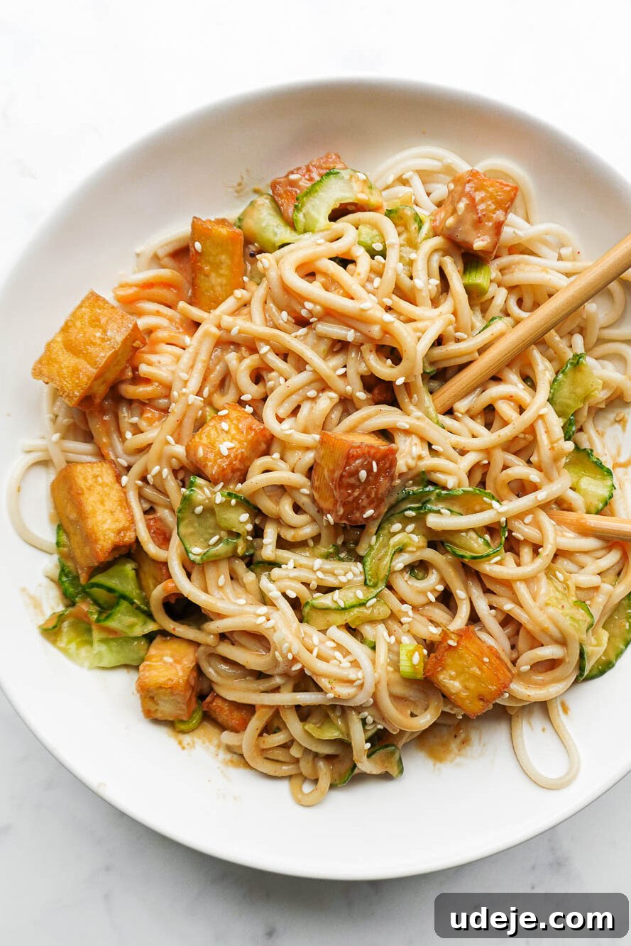 A healthy and visually appealing bowl of peanut noodles, garnished with crisp tofu, spiralized cucumber, chopped scallions, and sesame seeds, alongside a pair of chopsticks.