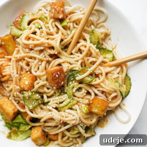 Bowl of peanut noodles topped with crispy tofu, spiralized cucumber, scallions, and sesame seeds with chopsticks