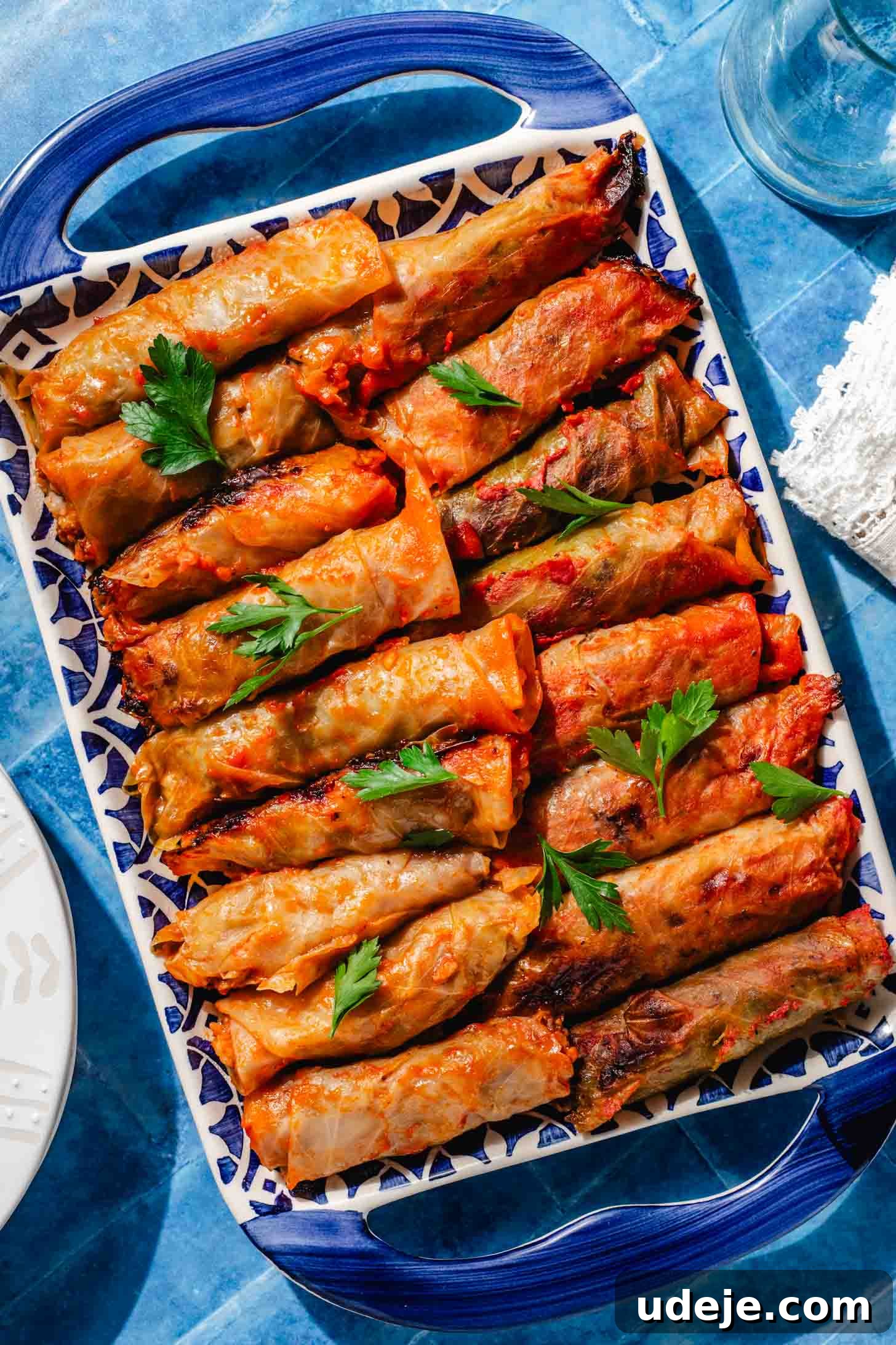 Irresistible Beef and Rice Cabbage Rolls 2 Perfectly baked cabbage rolls resting on a blue patterned tray, garnished with fresh parsley. The scene is set against a complementary blue background with a delicate white lace tea towel, a water glass, and white plates, suggesting a warm and inviting meal.