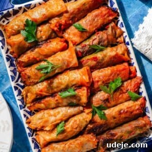 Cabbage rolls on a blue patterned tray garnished with parsley on a blue background with a white lace tea towel, a water glass and white plates at the side.