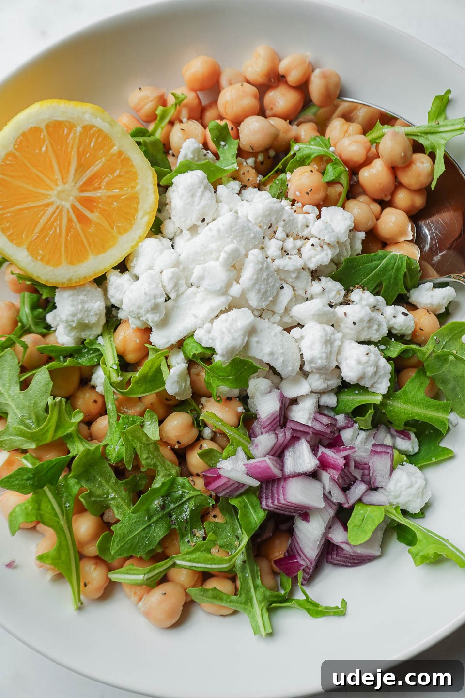 Vibrant Vegan Lemon Chickpea Salad 4 A vibrant bowl of chickpea feta salad ready for serving, garnished with fresh lemon wedges, finely diced red onion, bright green arugula, and golden chickpeas, highlighting the freshness of ingredients.