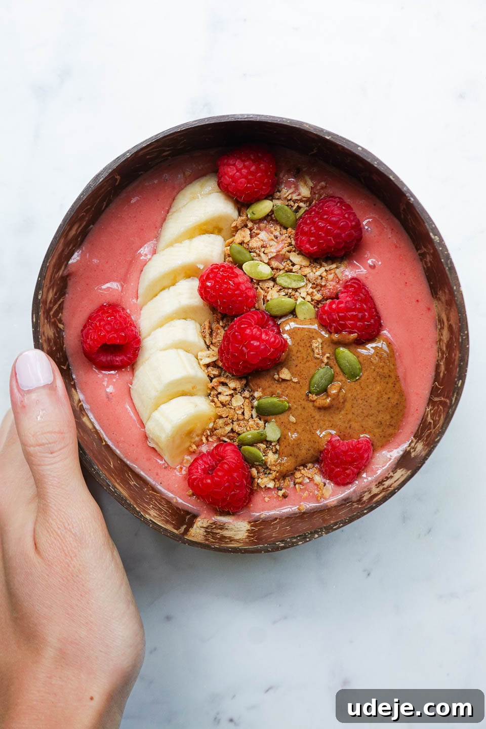 Simple and Delicious Vegan Raspberry Smoothie Bowl 2 hand holding a vegan raspberry smoothie bowl in a coconut bowl topped with granola, sliced banana, fresh raspberries, almond butter, pumpkin seeds