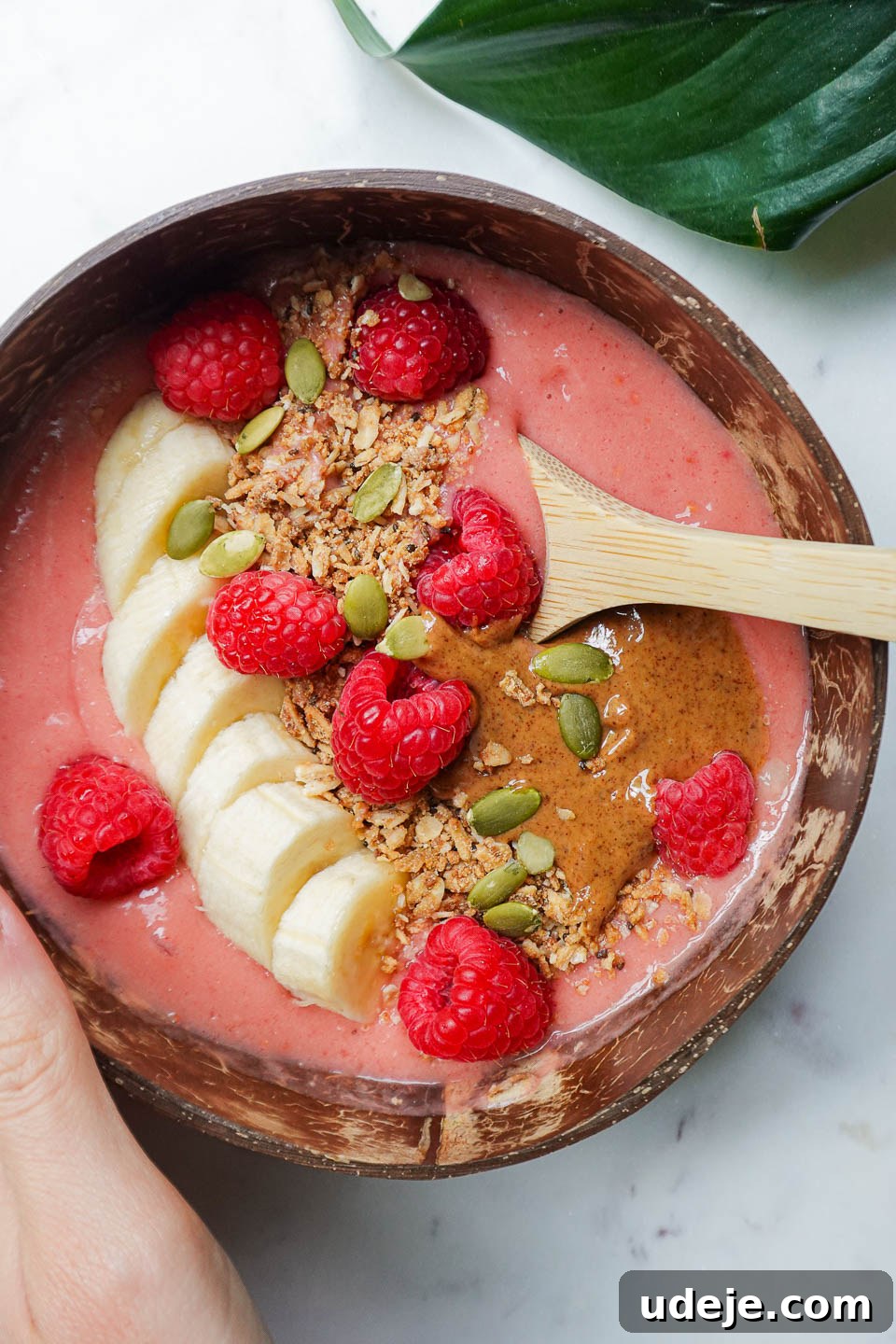 Simple and Delicious Vegan Raspberry Smoothie Bowl 3 hand holding a vegan raspberry smoothie bowl with a spoon inside, in a coconut bowl, topped with granola, sliced banana, fresh raspberries, almond butter, pumpkin seeds