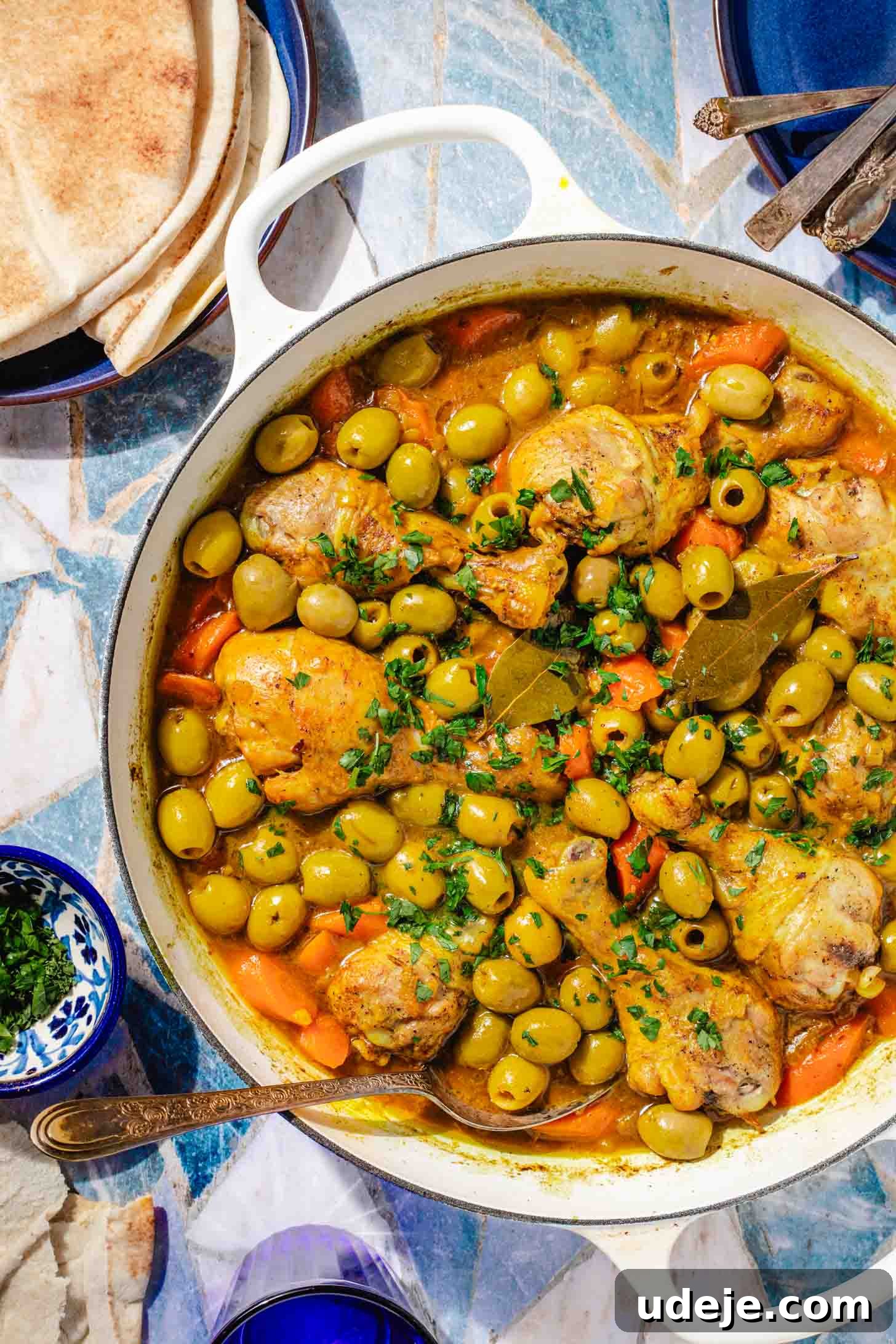 Chicken and Green Olive Tagine 2 A beautifully presented chicken stew with carrots, whole green olives, and bay leaves in a white pot, accompanied by a spoon. A blue plate with pita bread, another blue plate with spoon ends, and a small blue bowl of chopped parsley sit on a blue tile background, inviting viewers to savor this North African delight.