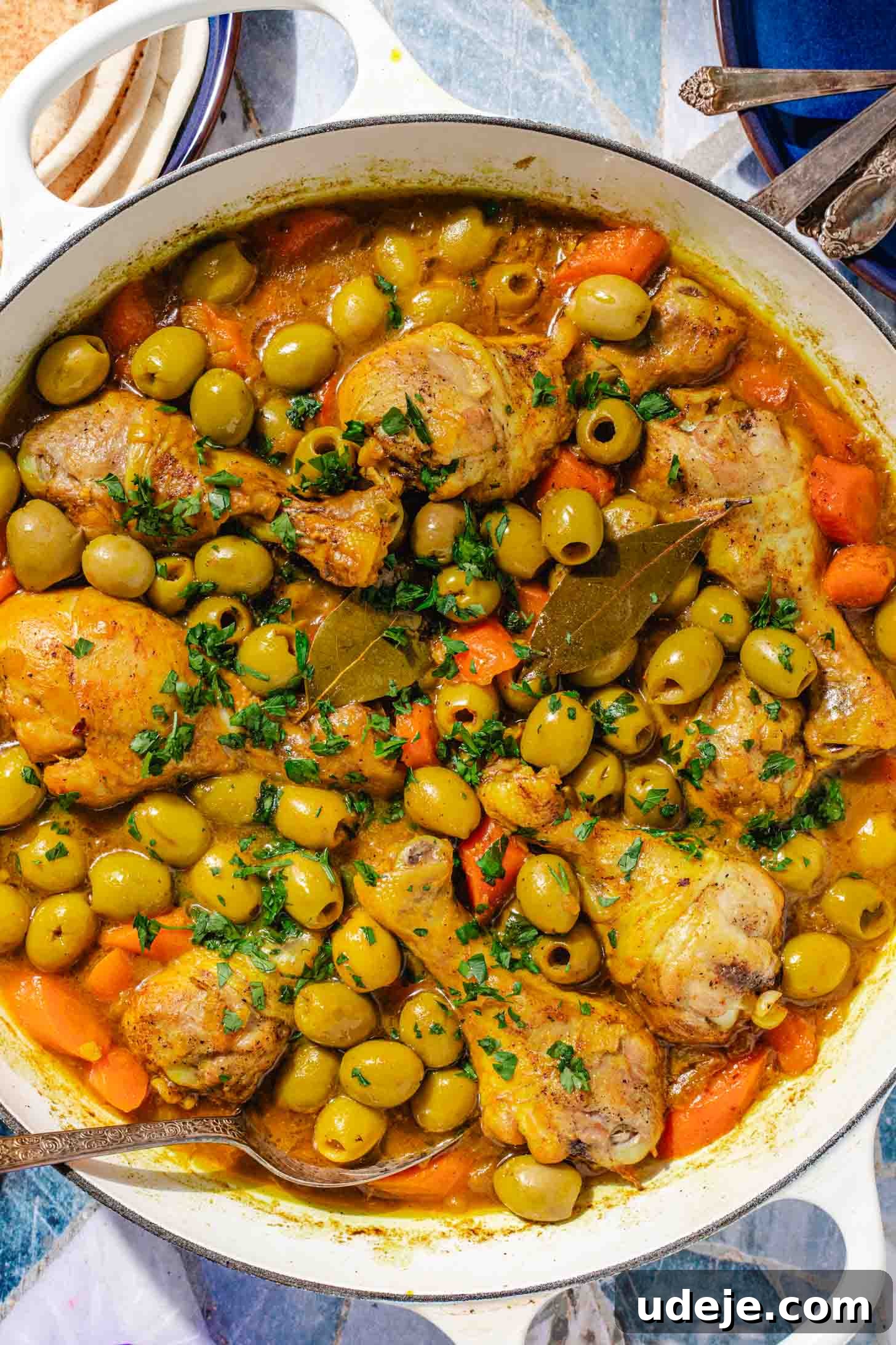 Chicken and Green Olive Tagine 12 A comforting serving of chicken stew with carrots, whole green olives, and bay leaves in a white pot with a spoon. A blue plate with pita bread and another blue plate with spoon ends are arranged on a blue tile background, inviting enjoyment.