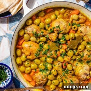 Chicken stew with carrots, whole green olives, bay leaves in a white pot with a spoon in it, a blue plate with pita, a blue pate with the ends of spoons, a small blue bowl with chopped parsley on a blue tile background.