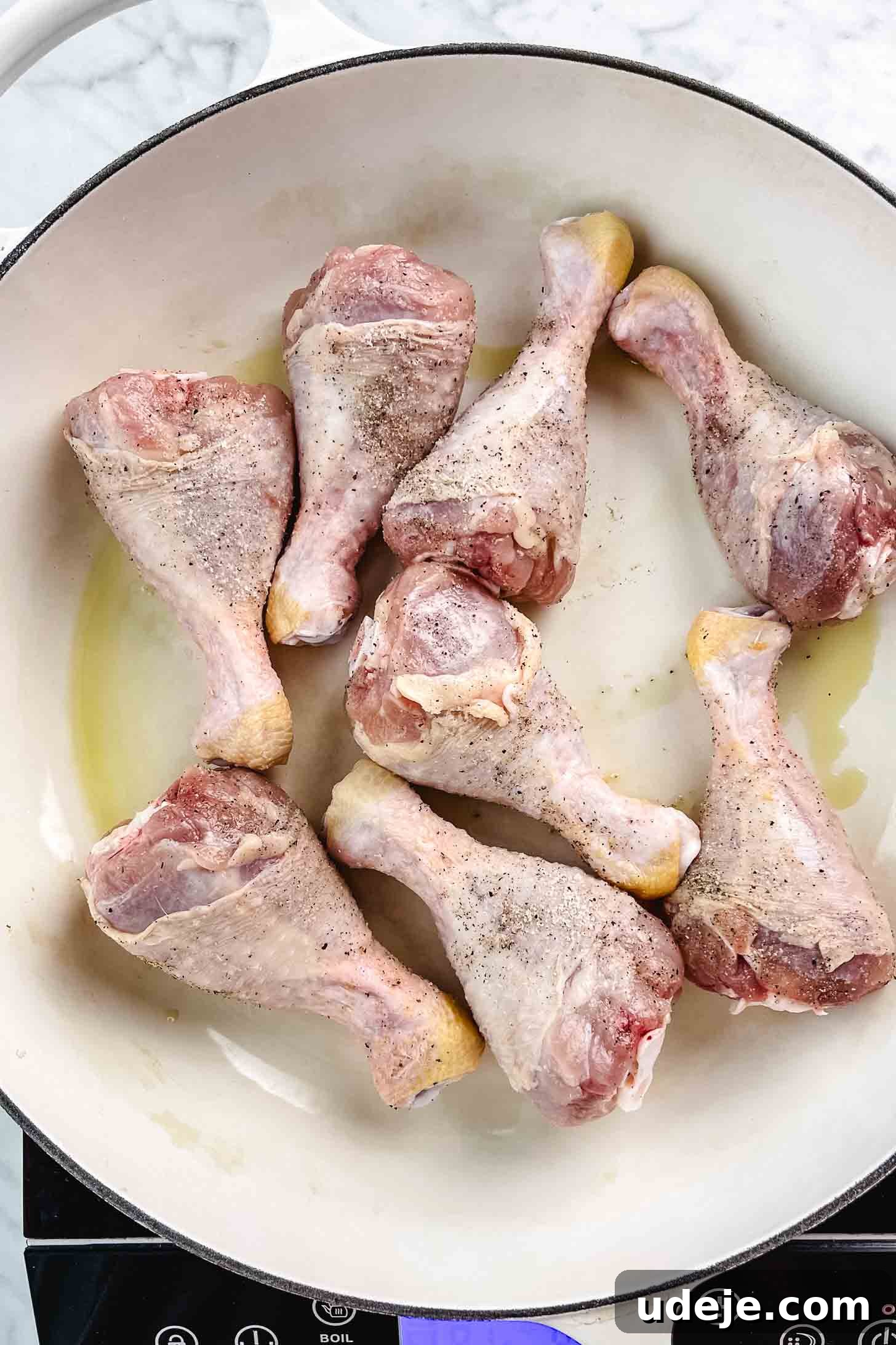 Chicken and Green Olive Tagine 4 Chicken legs seasoned with salt and pepper, starting to fry in olive oil in a large white pan on a marble background, creating a delicious base for the stew.