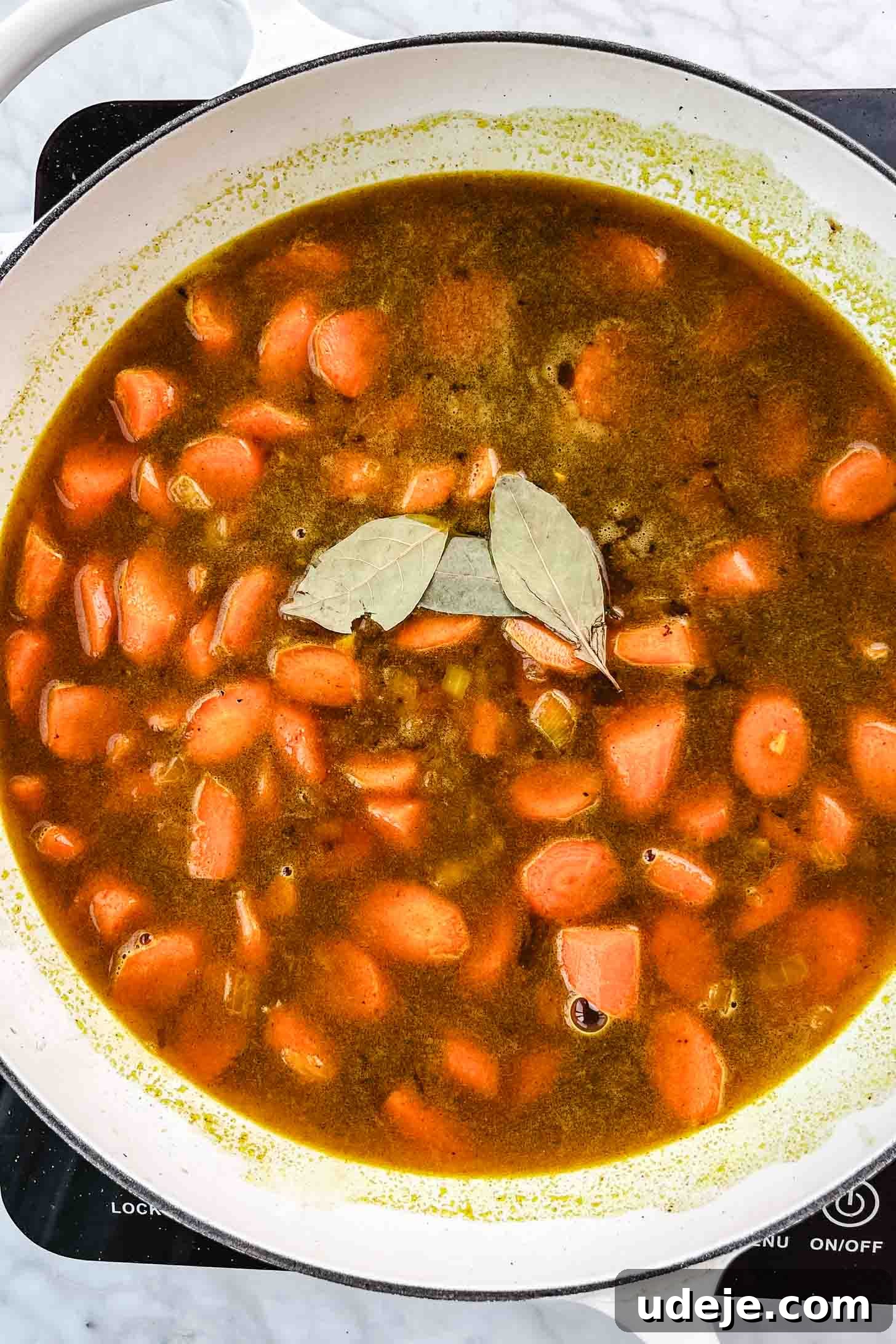 Chicken and Green Olive Tagine 9 Sliced carrots submerged in a rich, brown broth with bay leaves on top, simmering gently in a white pan on a marble background, allowing flavors to deepen.