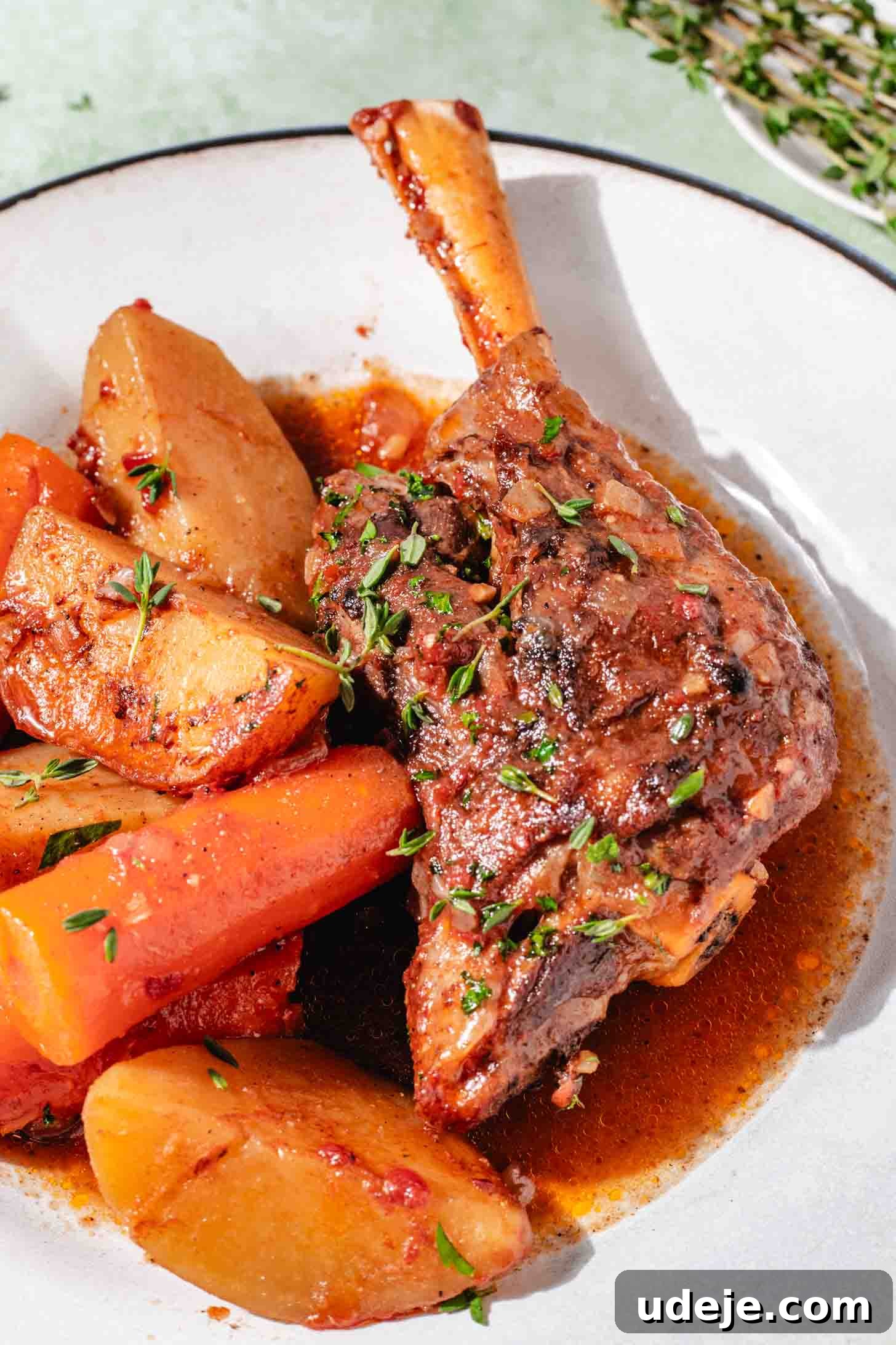 Melt-in-Your-Mouth Oven Braised Lamb Shanks 11 A single, perfectly braised lamb shank, boasting fall-off-the-bone tenderness, is elegantly plated alongside tender carrots and potato wedges, all glistening with the rich, aromatic broth. Fresh thyme sprigs provide a verdant contrast. The dish rests on a white plate with a subtle blue rim, set against a mottled green background for a rustic yet refined presentation.