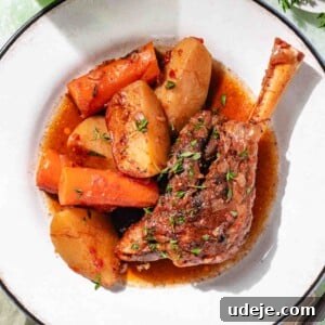 Lamb shank with broth, pieces of carrot and wedges of potato on a white blue rimmed plate with a green glass of water a small white plate with fresh thyme, a sage green tea towel on a mottled green background with a wooden ended knife and a metal fork at the side.