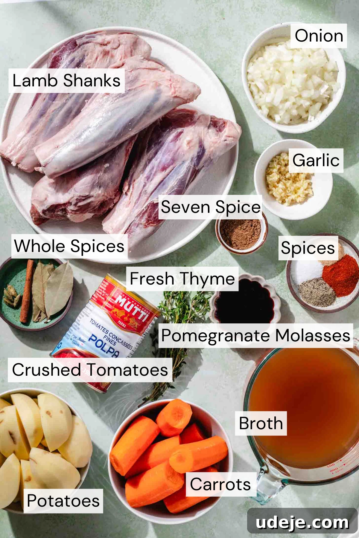 Melt-in-Your-Mouth Oven Braised Lamb Shanks 3 A top-down view of carefully arranged ingredients for Braised Lamb Shanks. Visible are four raw lamb shanks, whole spices like cinnamon sticks and cardamom pods, ground spices (seven-spice blend), fresh thyme sprigs, finely chopped onion and garlic, a can of crushed tomatoes, a bottle of dark pomegranate molasses, rich broth, and large pieces of sliced potatoes and carrots.
