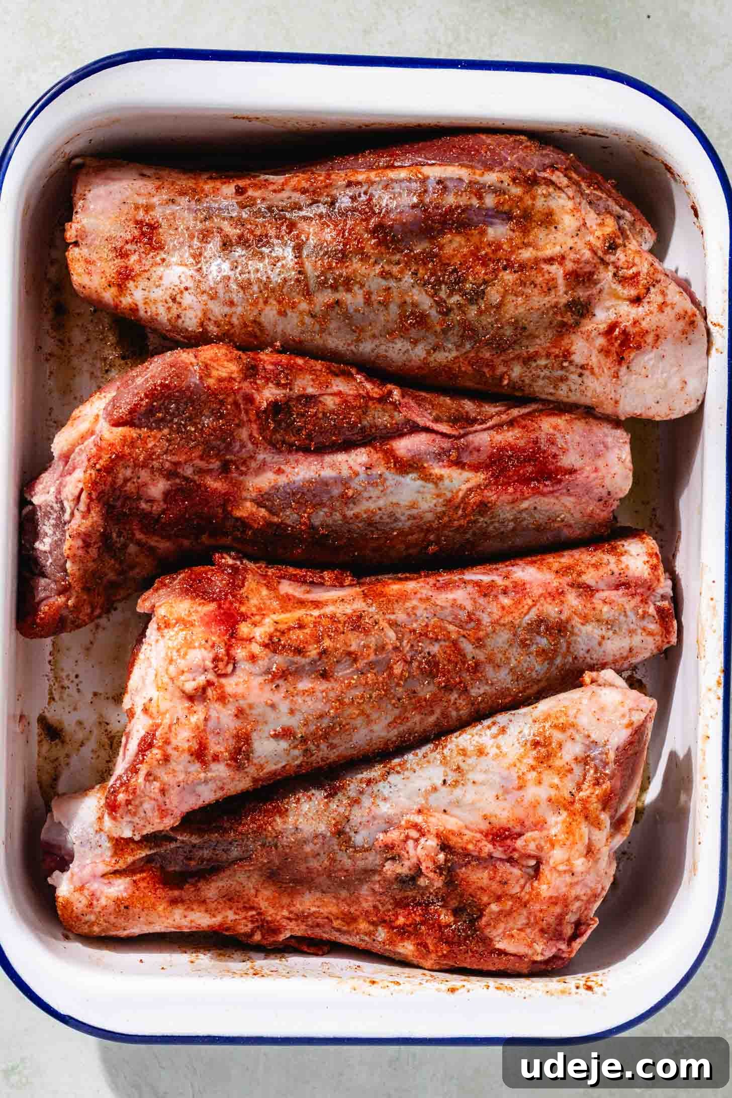 Melt-in-Your-Mouth Oven Braised Lamb Shanks 4 Raw lamb shanks generously coated with a reddish-orange seasoning of salt, pepper, paprika, and garlic powder, resting in a white and blue rectangular dish, ready for searing.