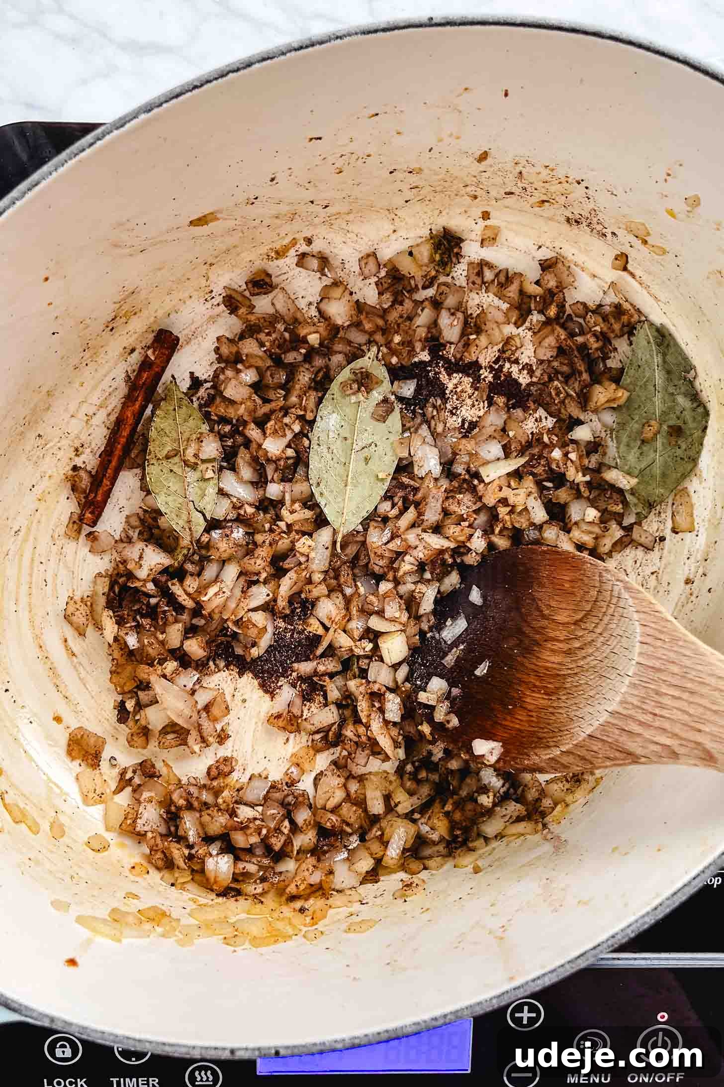 Melt-in-Your-Mouth Oven Braised Lamb Shanks 6 Diced yellow onions, softened and translucent, are sautéing in an oval Dutch oven with a cinnamon stick and bay leaves. A wooden spoon rests in the pot, stirring the aromatic mixture on a marble background.